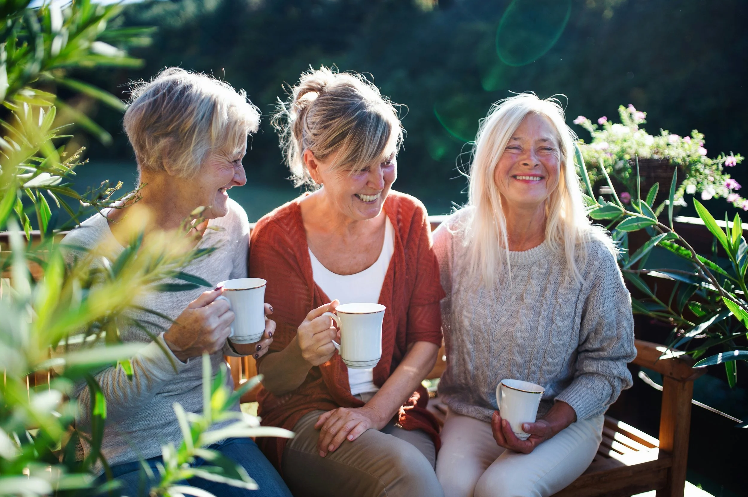 Four women are sitting outdoors on a wooden bench, smiling and holding white mugs, surrounded by greenery and potted flowers, enjoying a sunny day.