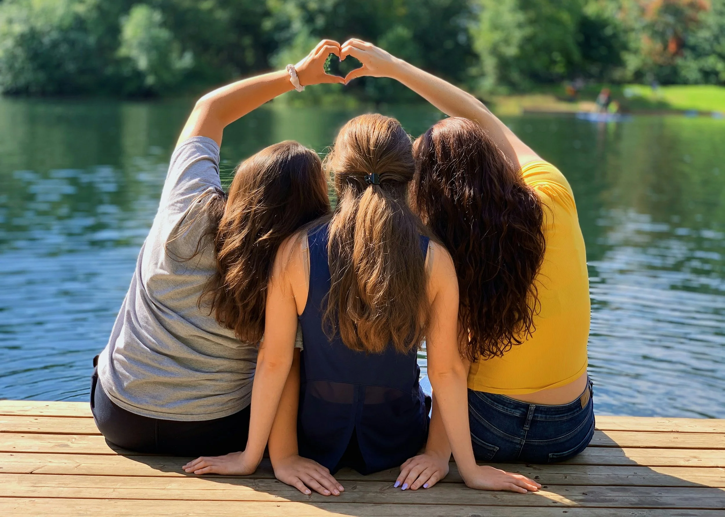 Three women sitting close together on wooden dock near water, facing away, and forming a heart shape with their hands.