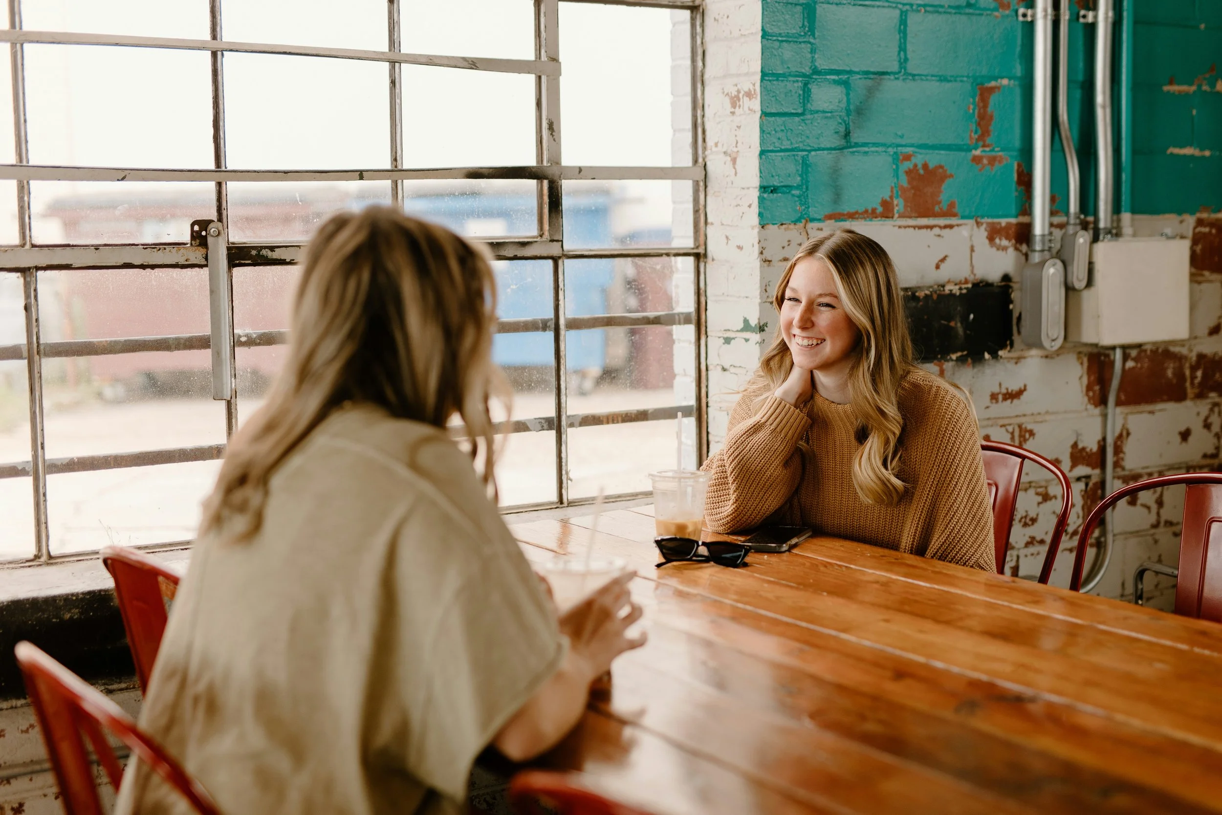 Two women sitting at a wooden table in a cafe, smiling and talking. One has long blonde hair and is wearing a beige sweatshirt, the other has wavy light brown hair and is wearing a brown sweater. A glass of iced coffee and sunglasses are on the table. The cafe has large windows with metal grids and a painted brick wall.