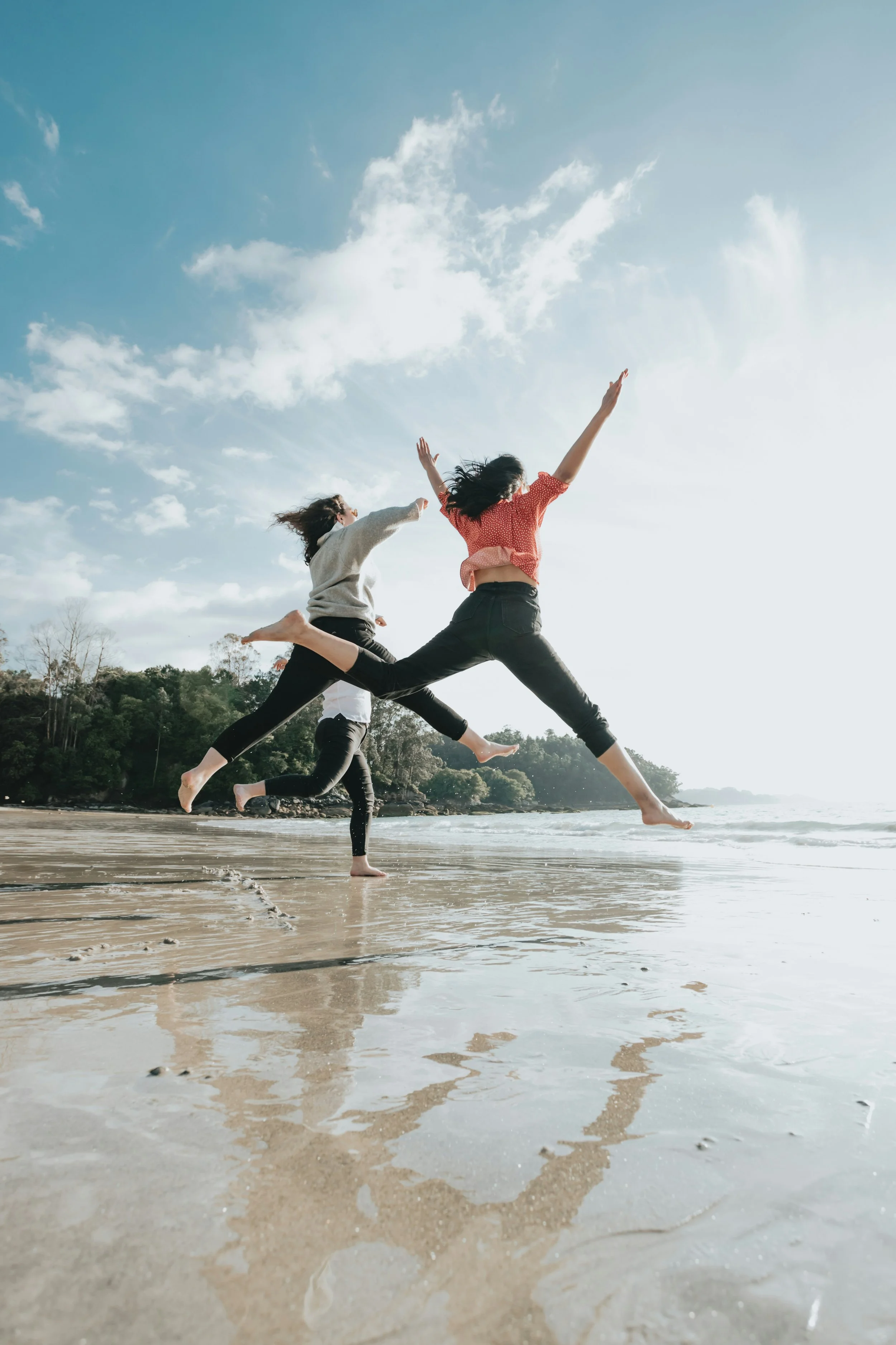 Three women jumping in the air on a sandy beach, with a backdrop of trees and a blue sky with clouds.