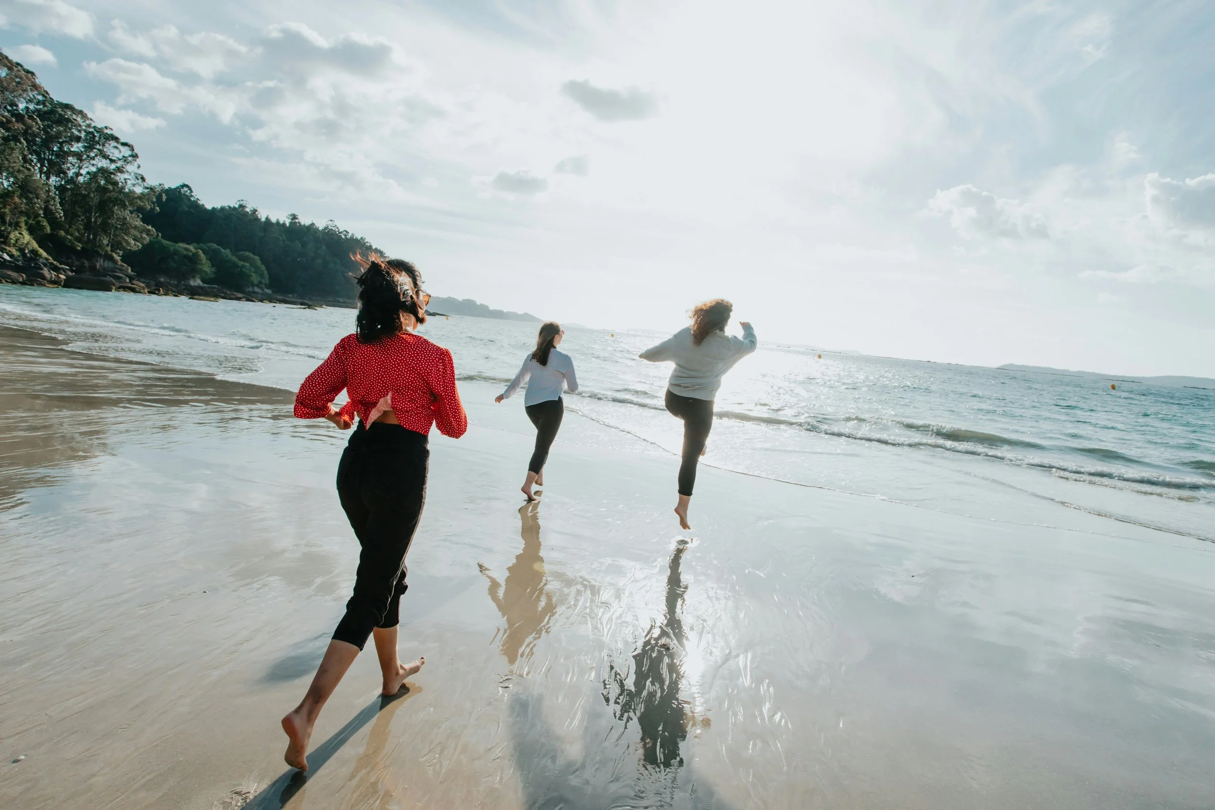 Three women walking along the beach with waves touching their feet, with a lush green hill in the background and clouds in the sky.
