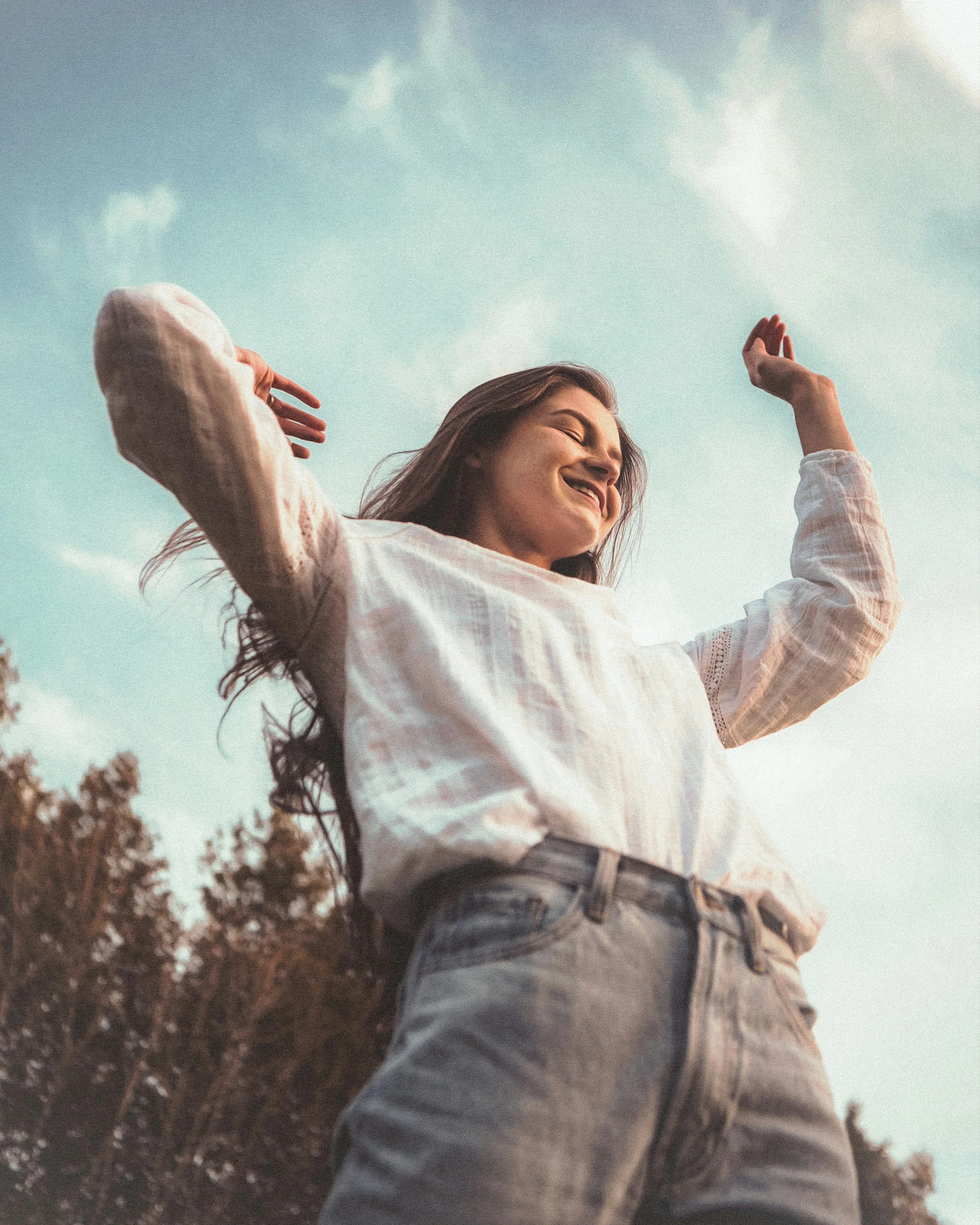 A young woman with long hair smiling, standing outdoors with a clear blue sky and some clouds overhead. She is wearing a light-colored long-sleeve top and jeans, with her hands raised near her head, enjoying a sunny day.