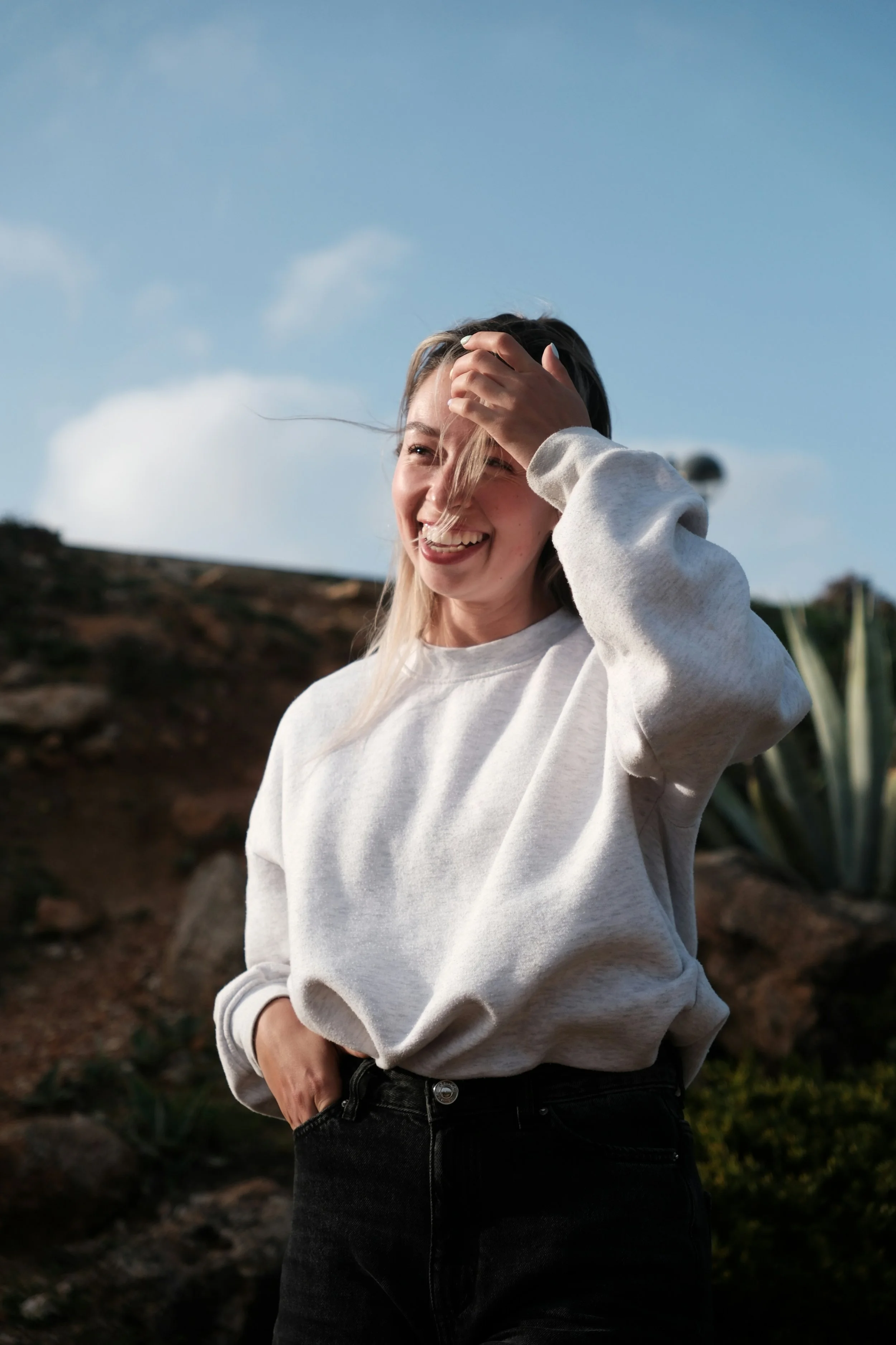 A young woman in a white sweatshirt and black pants standing outdoors on rocky terrain with a clear blue sky in the background. She is smiling, with her hand on her forehead, and appears happy.