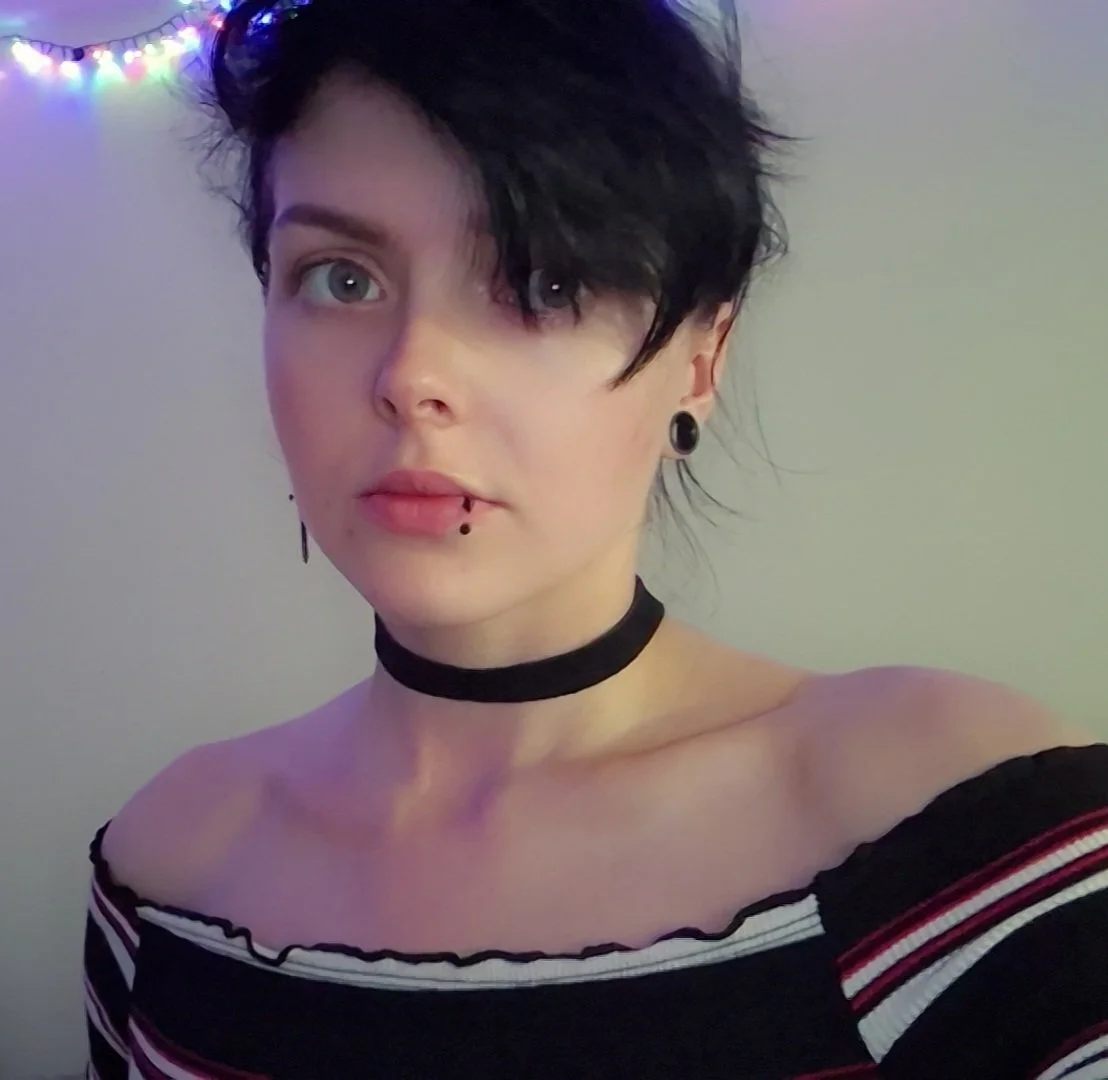 A young woman with short, black hair styled in a messy, side-swept manner looks at the camera. She has facial piercings, a piercing on her upper lip, and wears black plug earrings. She is wearing an off-the-shoulder striped top with black, white, and red horizontal stripes, and a black choker. In the background, there are colorful string lights hanging on a wall.