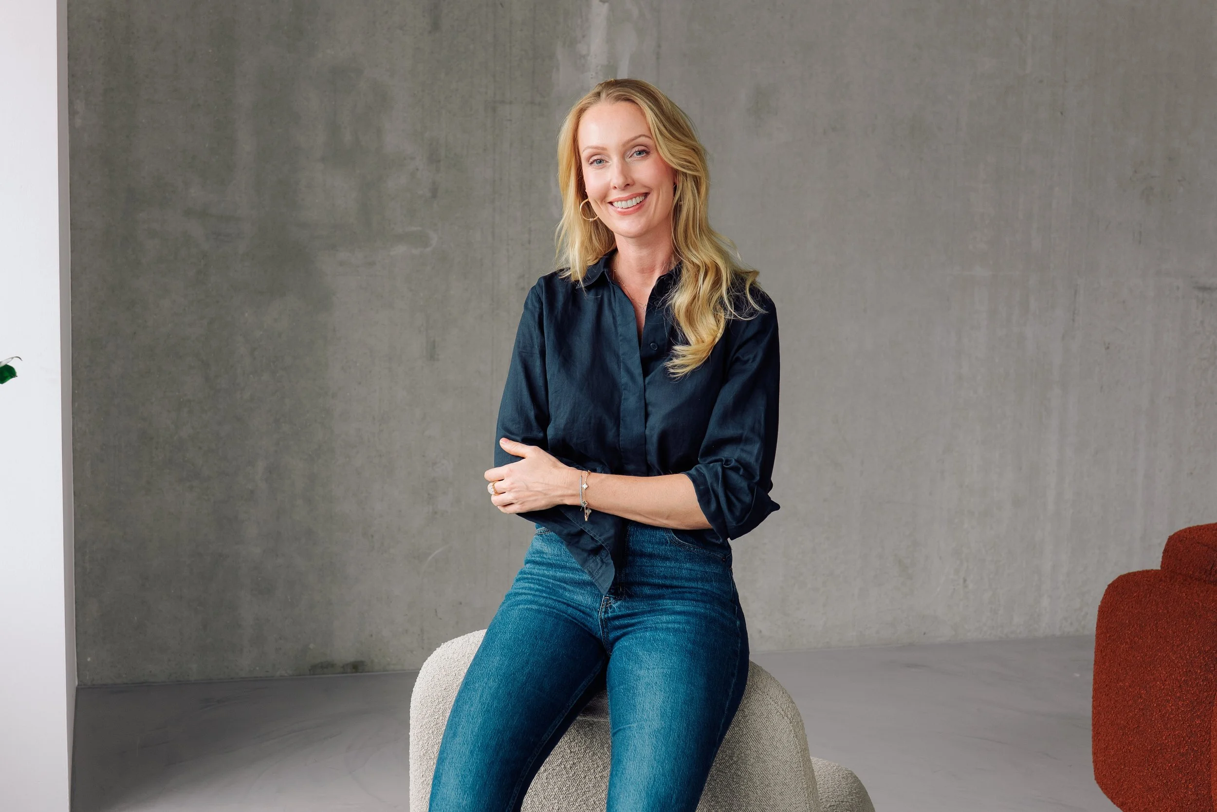 Eine lächelnde blonde Frau in einer schwarzen Bluse und blauen Jeans sitzt auf einem beigen Sitzmöbel vor einer grauen Wand.