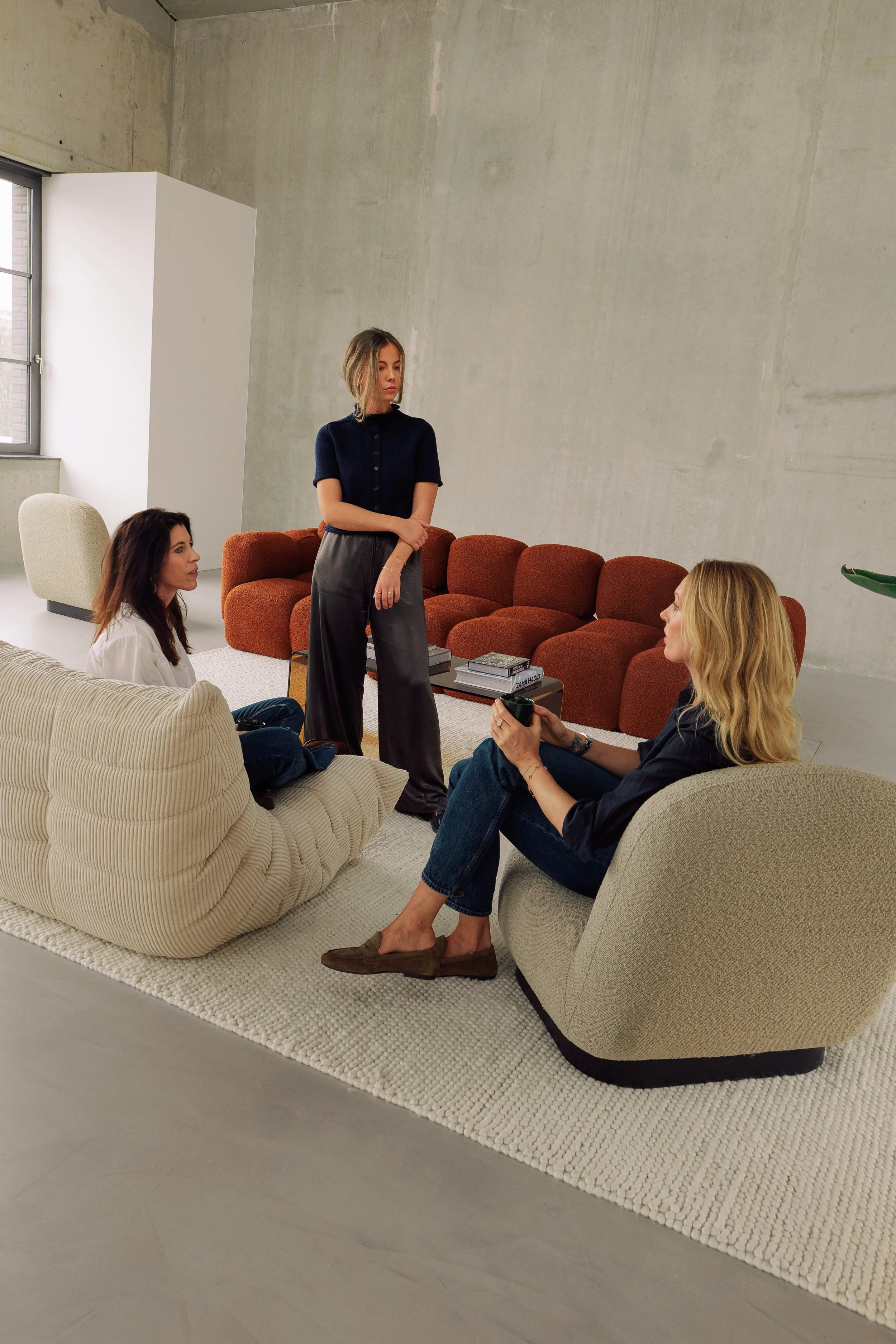 Vier Frauen sitzen und stehen in einem modernen Wohnzimmer, unterhalten sich und trinken Kaffee, auf einem weißen Teppich vor einem roten Sofa, mit Bücher und einem Pflanzen im Hintergrund.