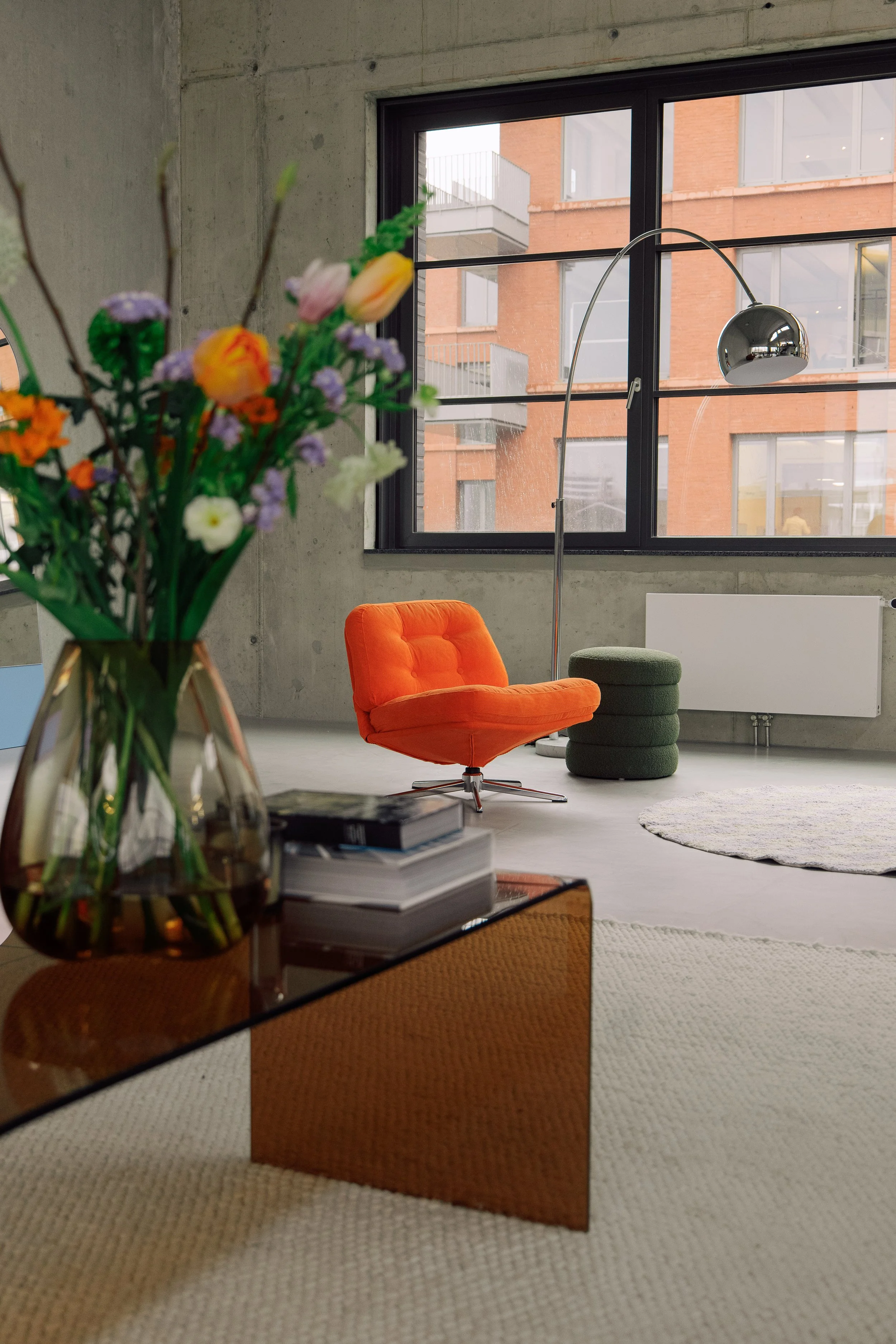 Innenaufnahme eines modernen, minimalistischen Wohnzimmers mit großen Fenstern, einem orangefarbenen Sessel, einem grünen Podest, einem weißen Teppich und einer Stehlampe. Im Vordergrund befindet sich eine Vase mit bunten Blumen auf einem Tisch.