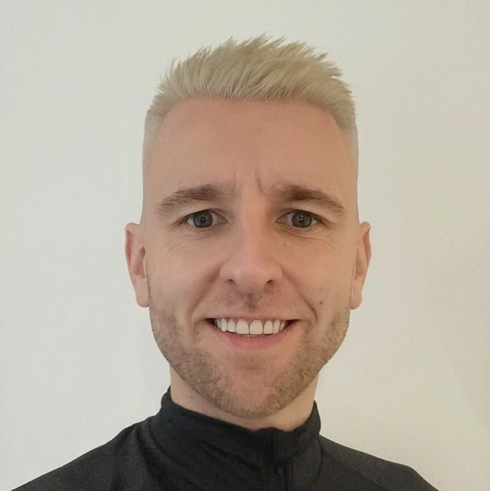 A young man with short blond hair, blue eyes, and light facial hair, smiling and wearing a black high-collared shirt, against a plain white background.