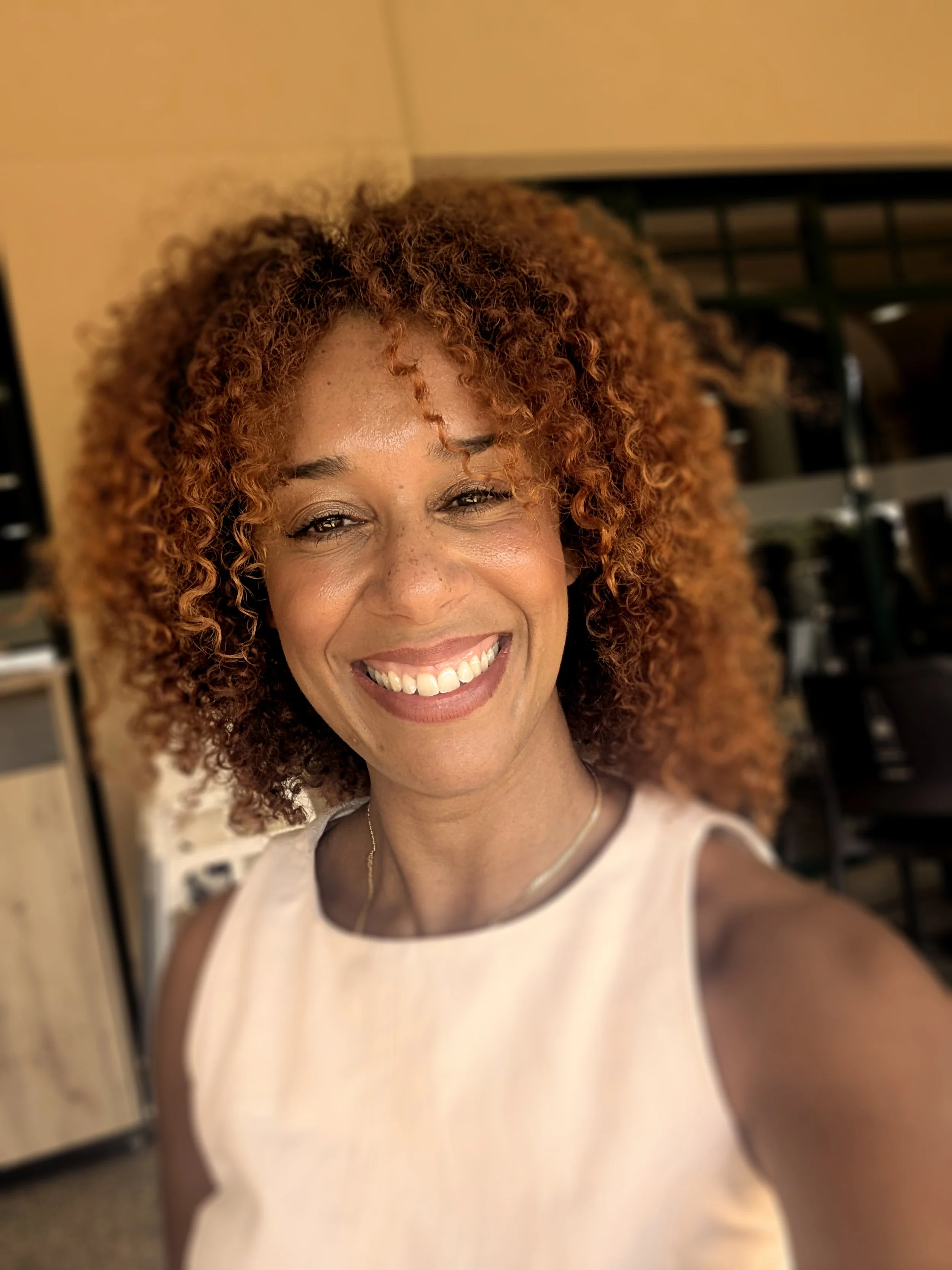 A woman with curly, reddish-brown hair smiling and looking at the camera in an indoor setting.