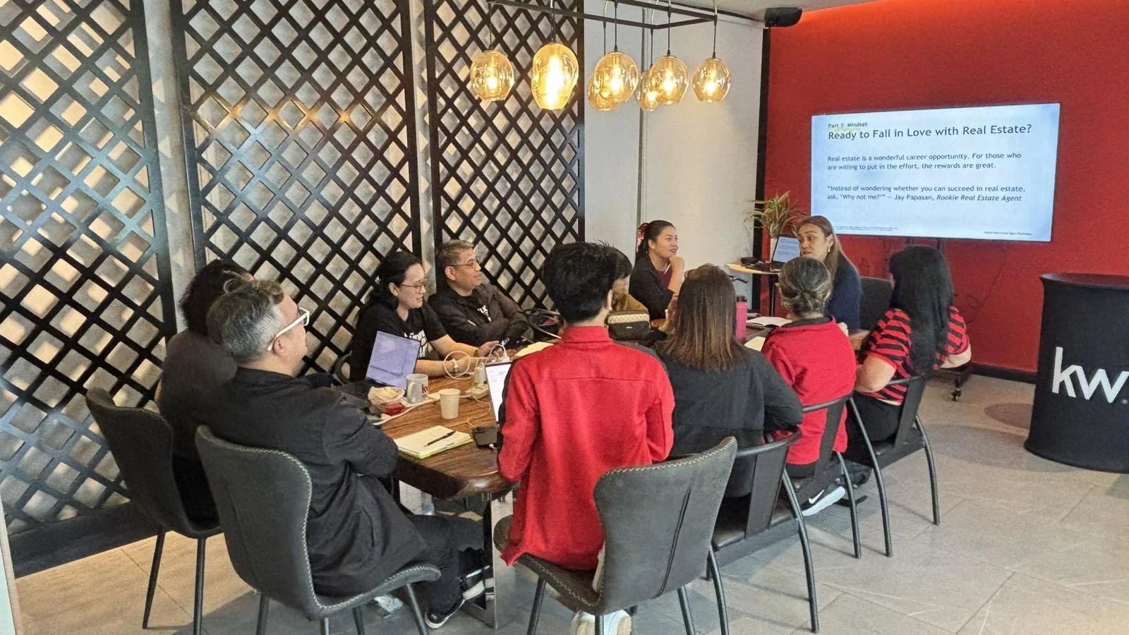 A group of people attending a presentation in a conference room. The room has a large screen displaying a slide about real estate. The attendees are seated around a table, some with laptops and notebooks. The decor includes hanging lights and a black lattice wall.