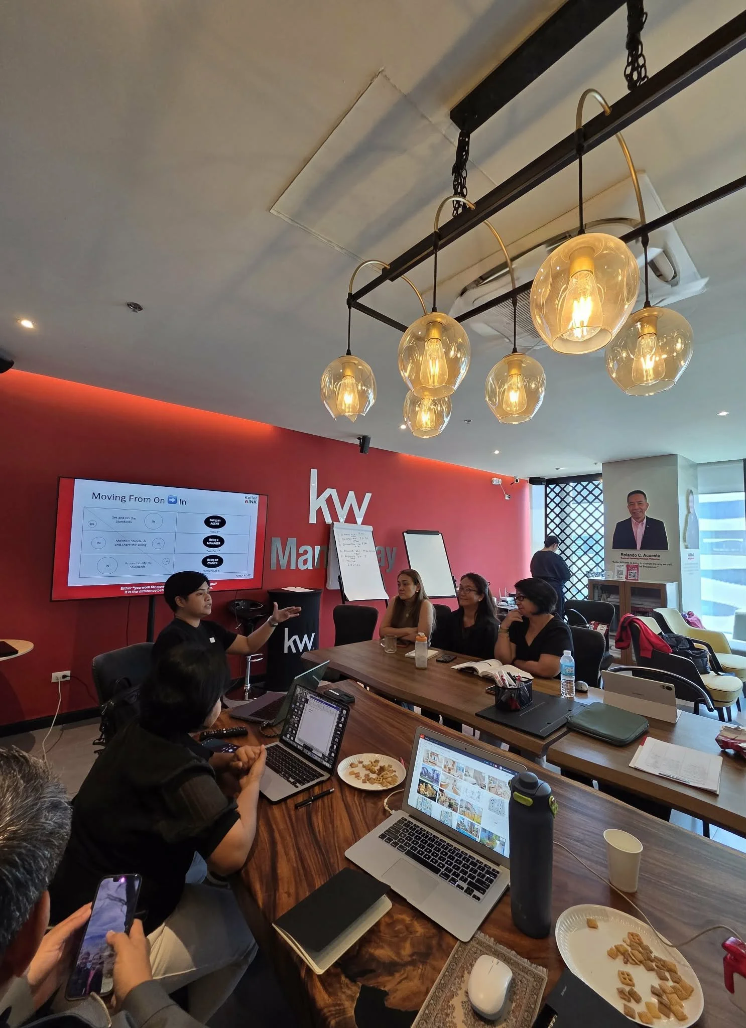 A group of people attending a presentation in a conference room with a red wall displaying the KW logo. A presenter is speaking, and there is a large screen showing a speaker. The room has modern hanging lights and several tables with laptops, notebooks, drinks, and snacks.
