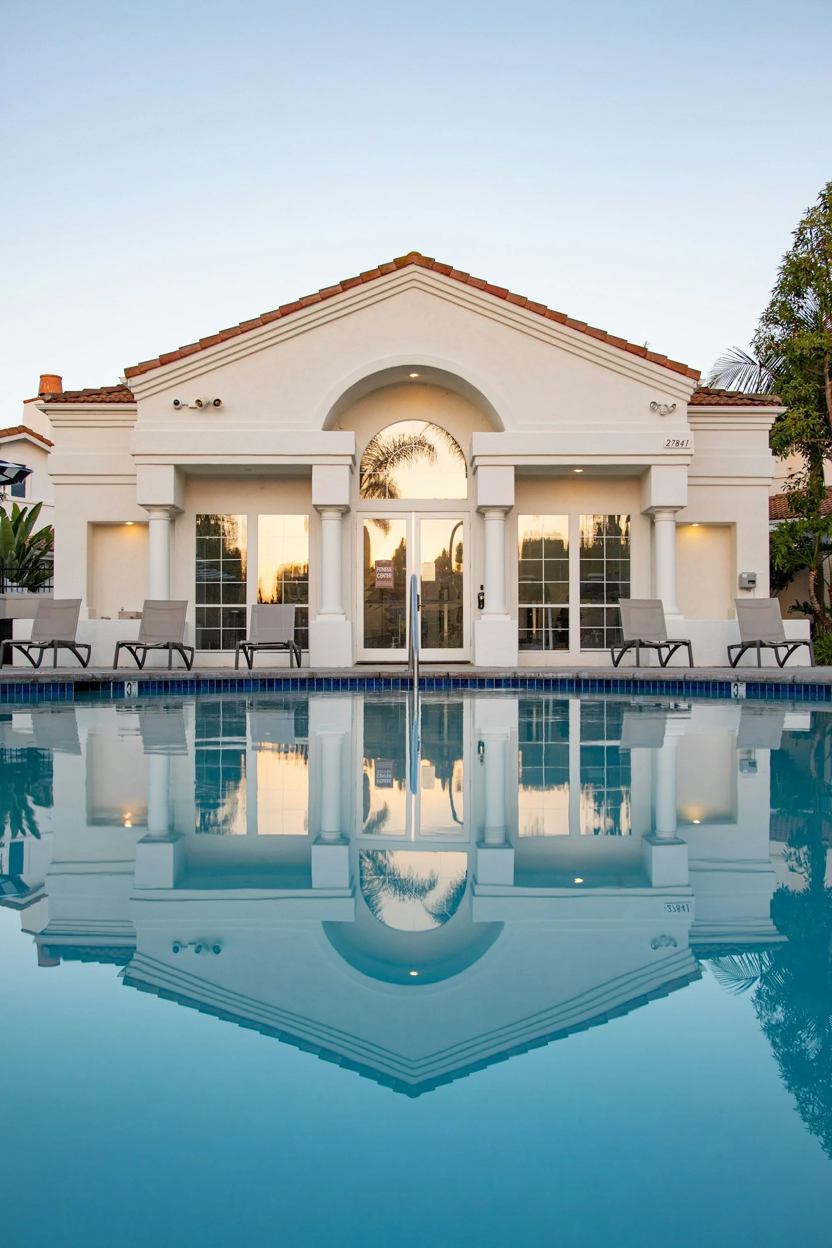 Front view of a white building with columns and a tiled roof, reflected in a swimming pool.