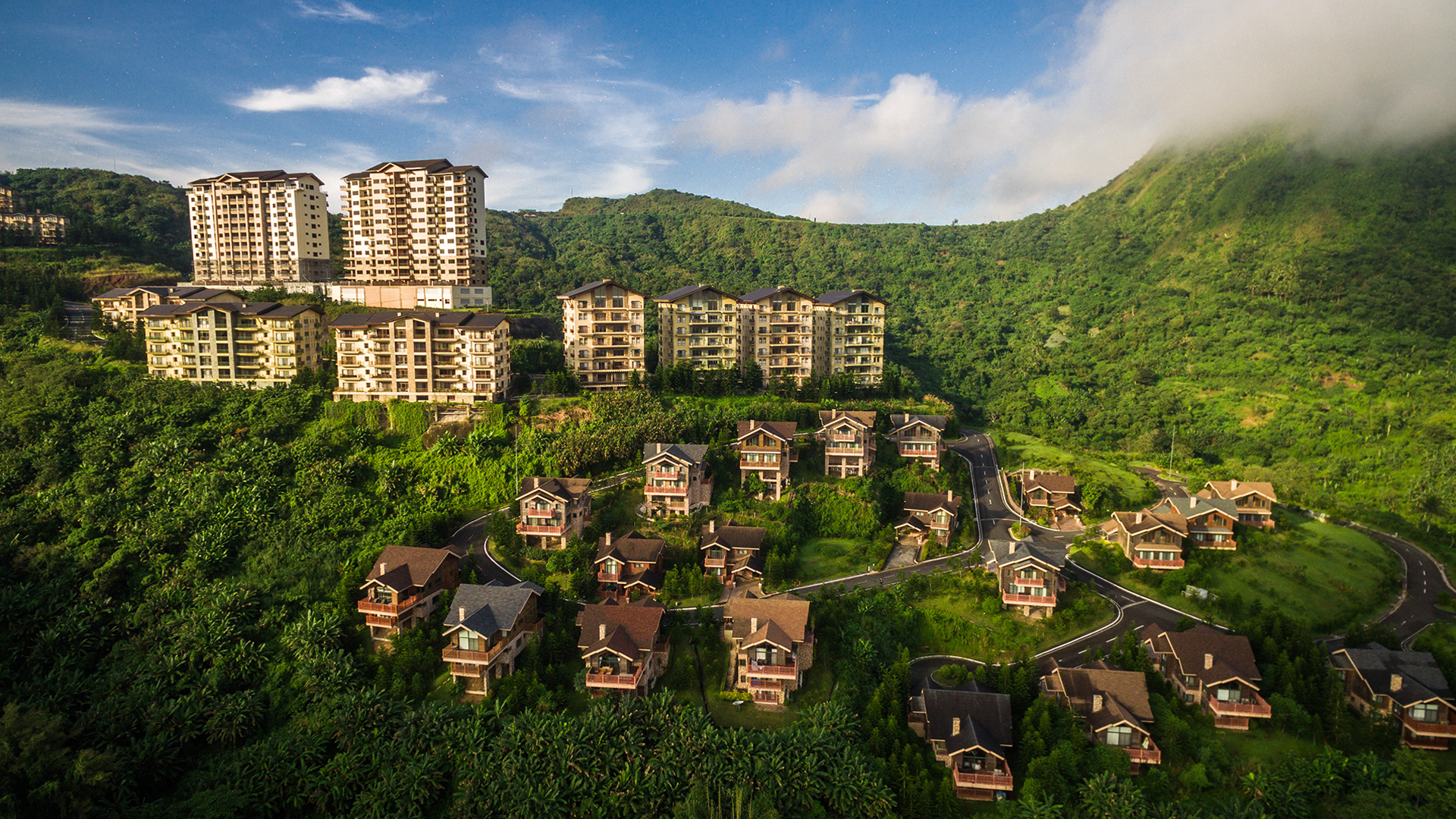 A hillside with multiple residential buildings, including high-rise apartments and smaller houses, surrounded by lush greenery and trees under a partly cloudy sky.