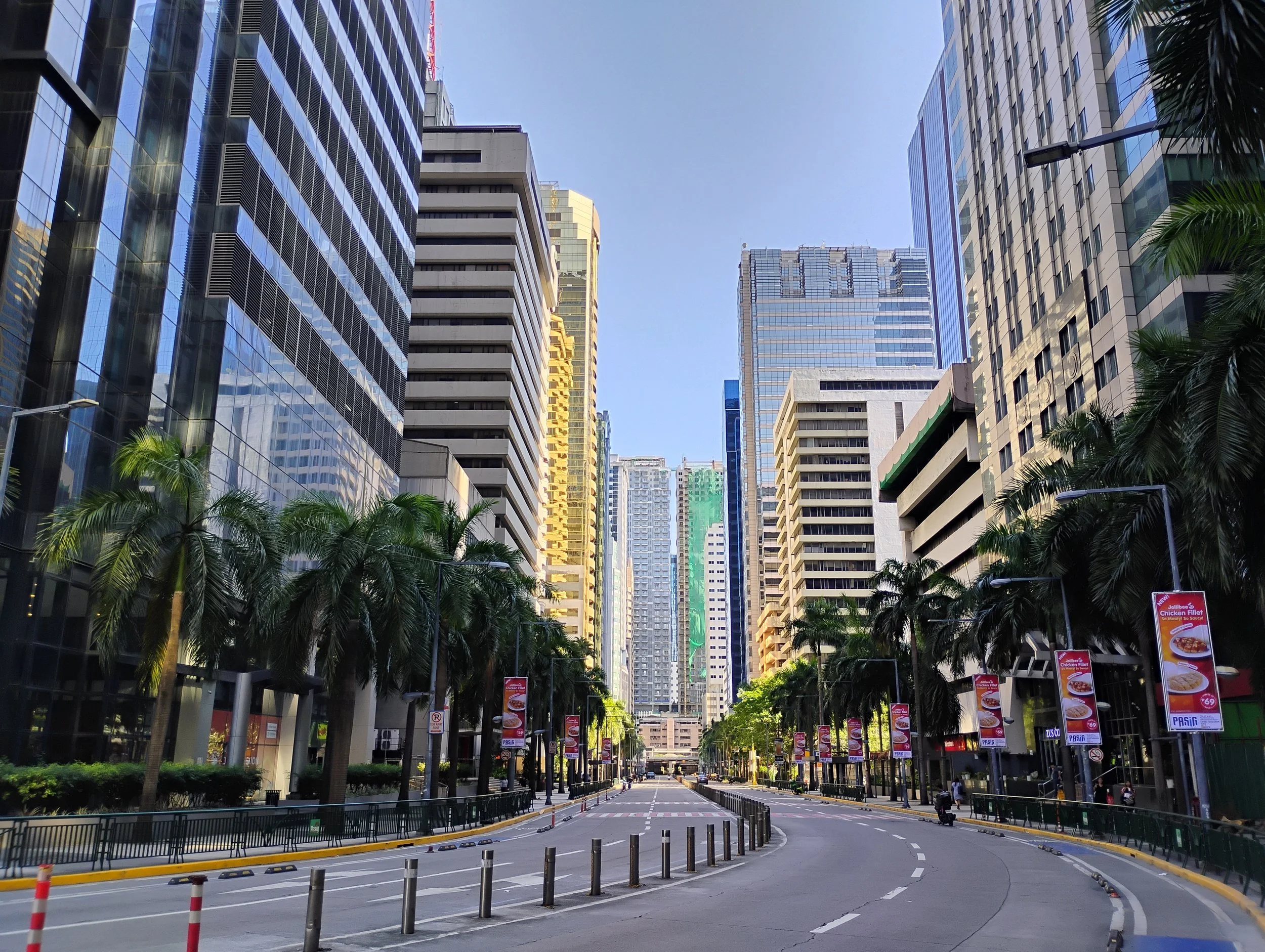 City street lined with tall buildings and palm trees, with a few pedestrians and vehicles, under a clear blue sky.
