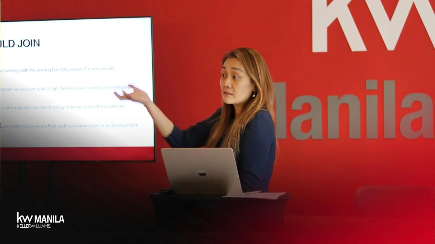 A woman with long hair presenting in front of a large screen, with a red wall behind her that has the word "Manila" in grey letters and a partial logo. She is pointing at the screen and has a laptop in front of her.