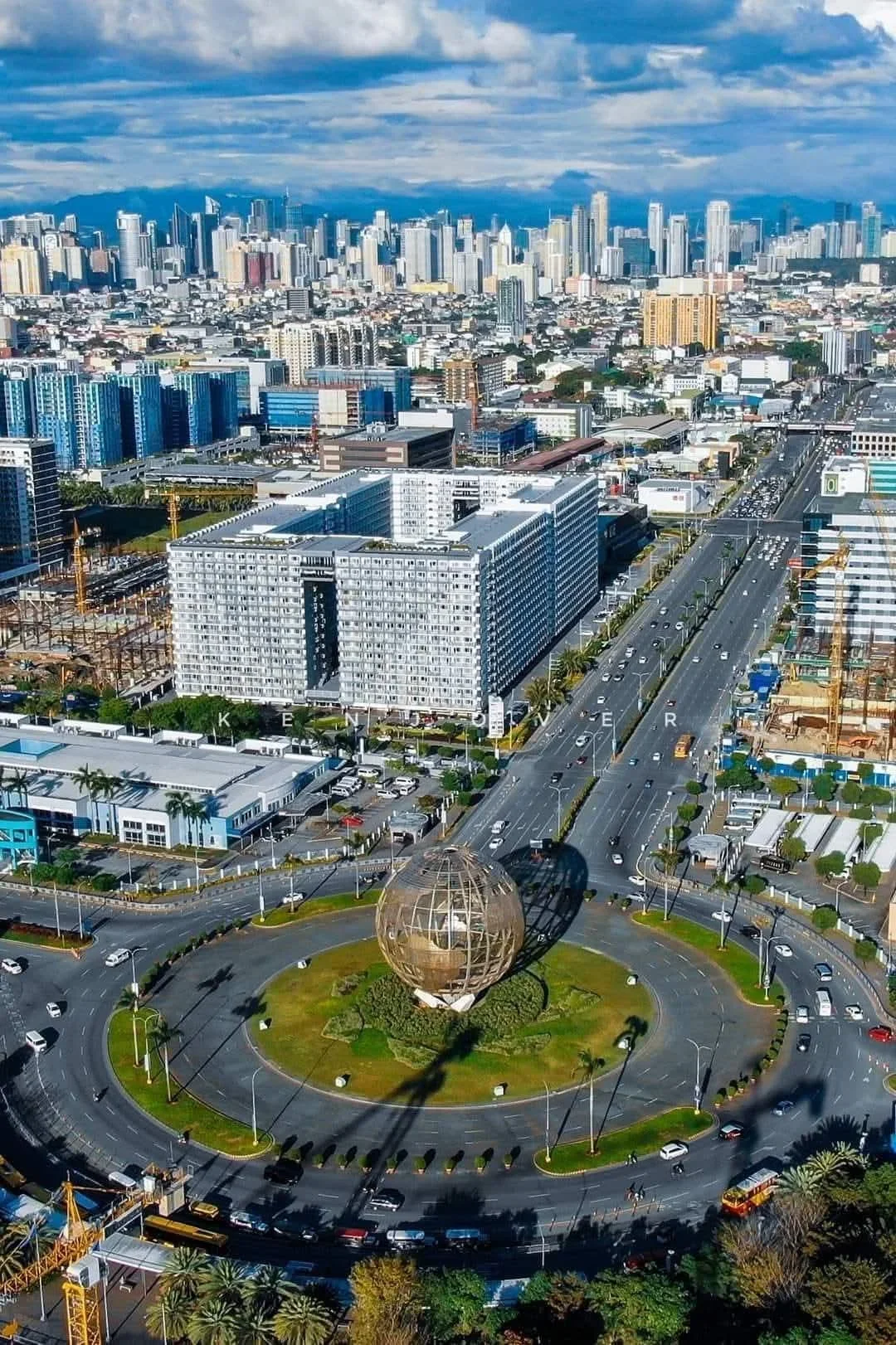Aerial view of downtown Los Angeles showing a circular park with a metallic globe sculpture, surrounded by roads filled with cars, and tall buildings in the background under cloudy skies.