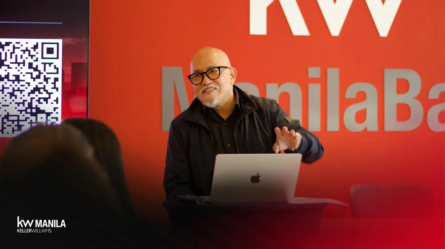 A man with glasses and a beard, wearing a black jacket, is speaking and gesturing with his right hand behind a silver MacBook. He is standing in front of a red background with large white and grey text that says "MW Manila" and "KellerWilliams." A QR code is visible on the left side of the background.