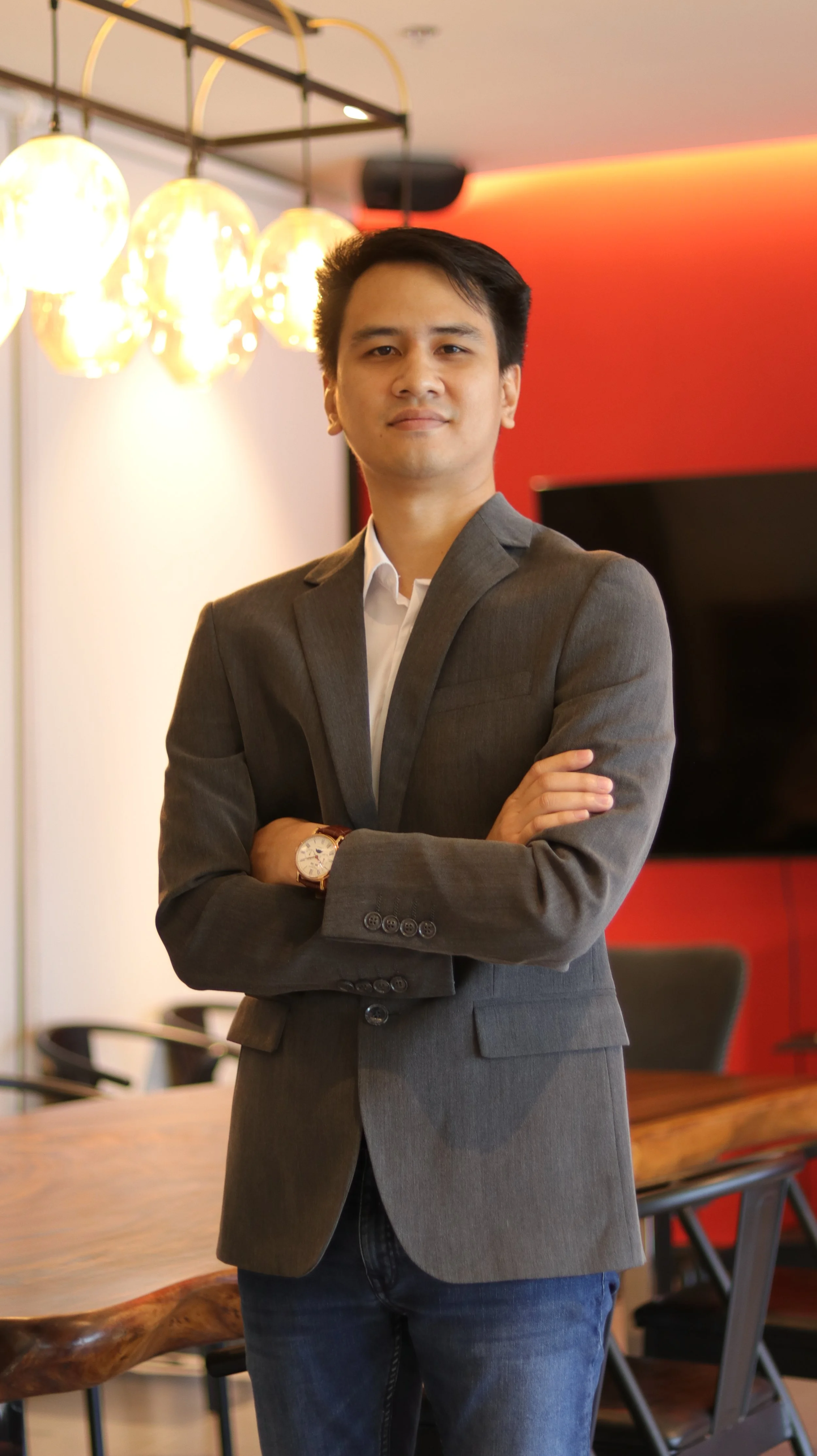 A confident Asian man in a gray suit and white shirt stands with arms crossed in a modern office or conference room with warm lighting, a red accent wall, and a large flat-screen TV.