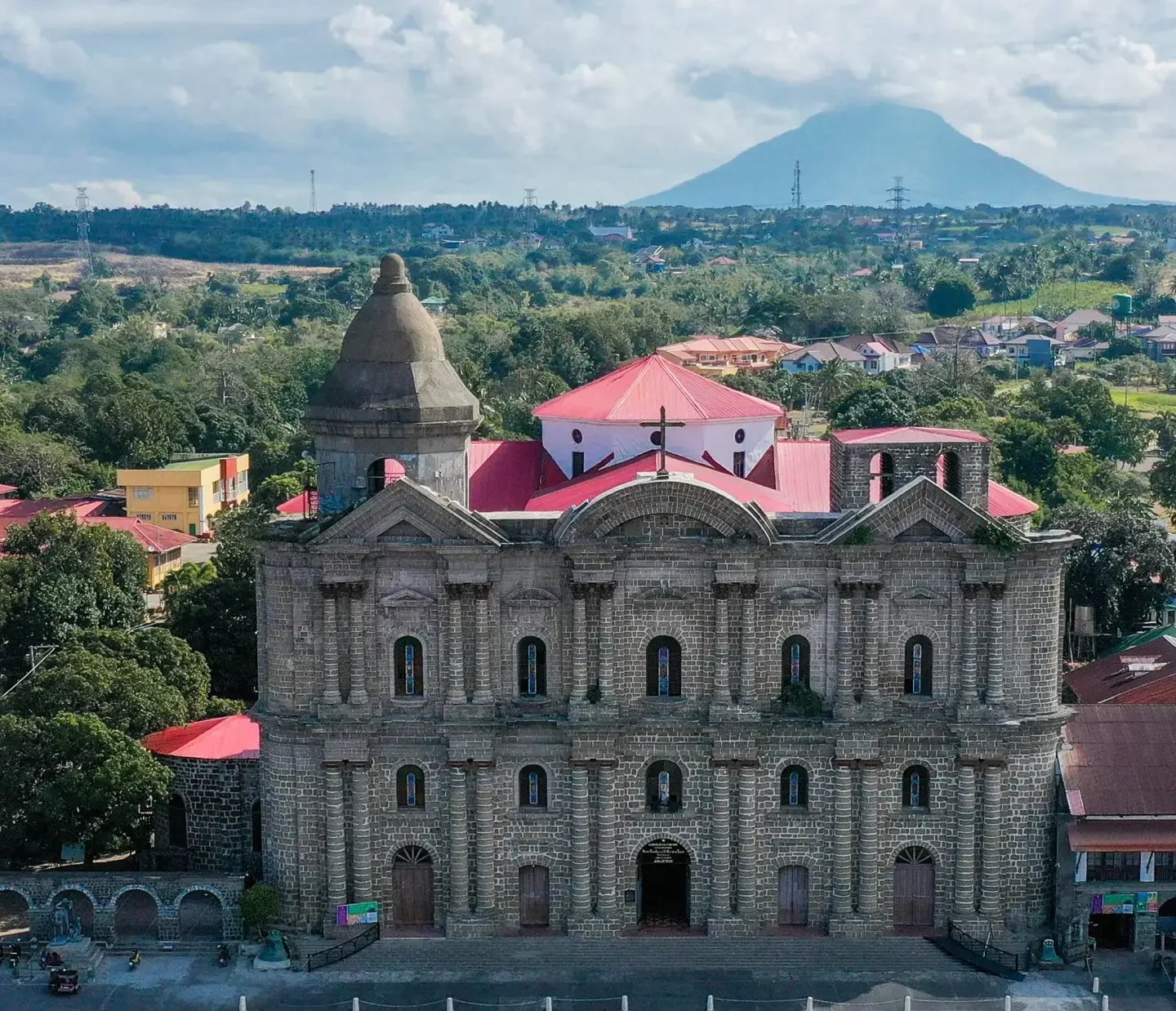 A historic stone church with a dome and tall bell tower, surrounded by green trees, residential houses with red and blue roofs, and a mountain in the background under a partly cloudy sky.