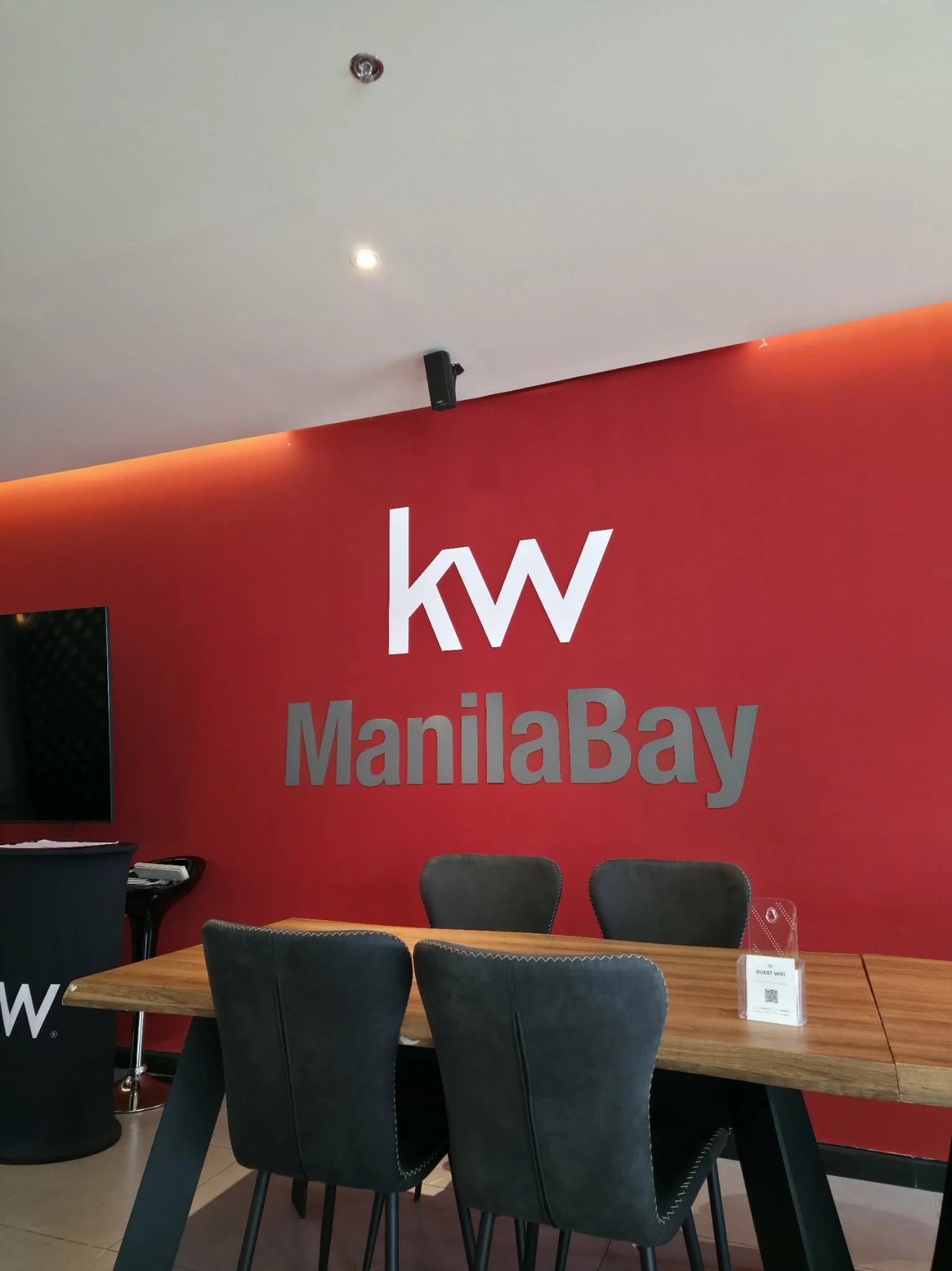 Interior of a real estate office with a red wall displaying the KW Manila Bay logo, a wooden table with four black chairs, a TV on the wall, and a small sign with a QR code on the table.