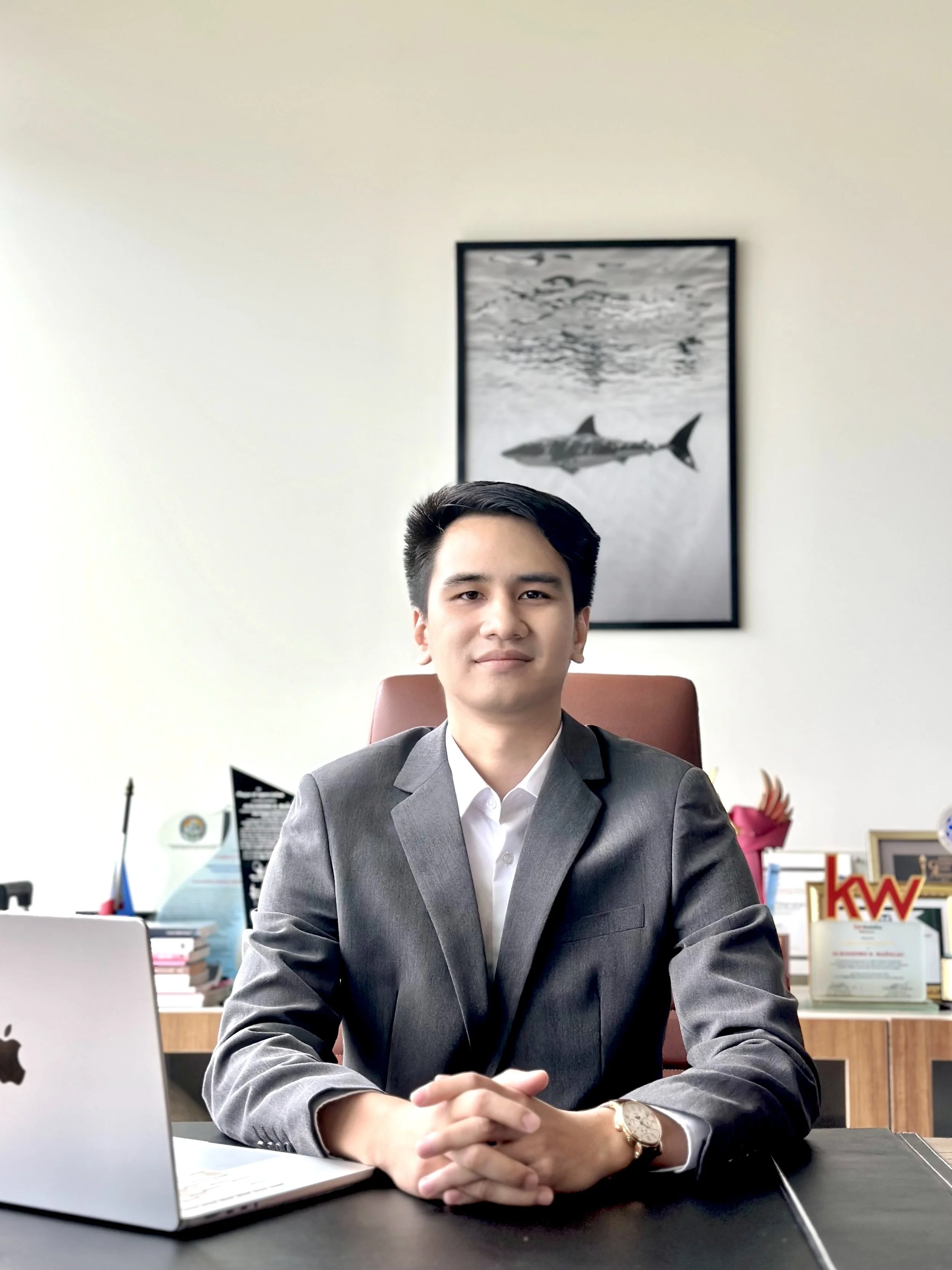 A young man in a gray suit sitting at a desk with hands clasped, in an office with a framed shark picture on the wall behind him and various office items on the desk and shelves.