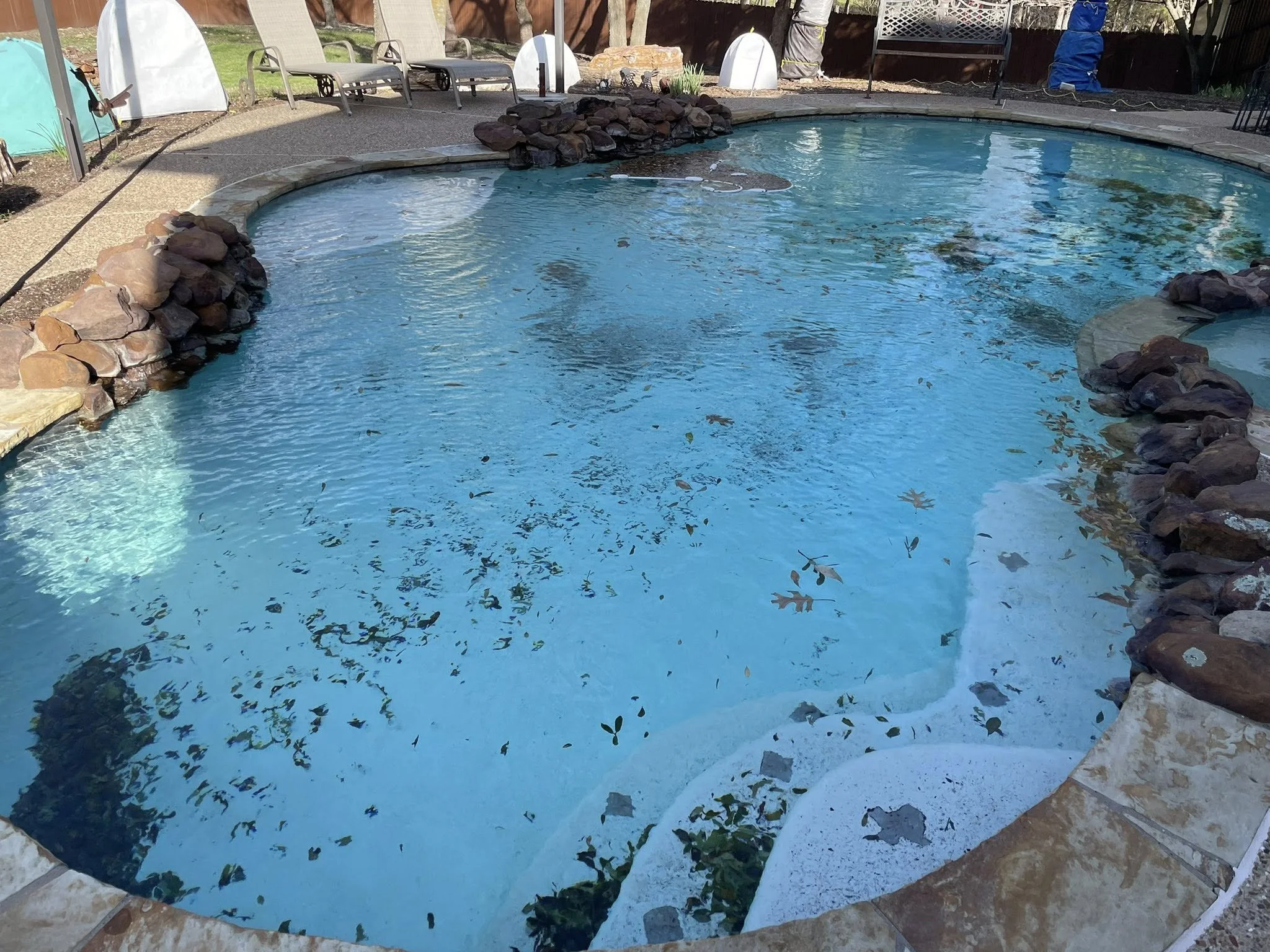 Backyard swimming pool with rocks lining the edges, surrounded by patio furniture and deck chairs.