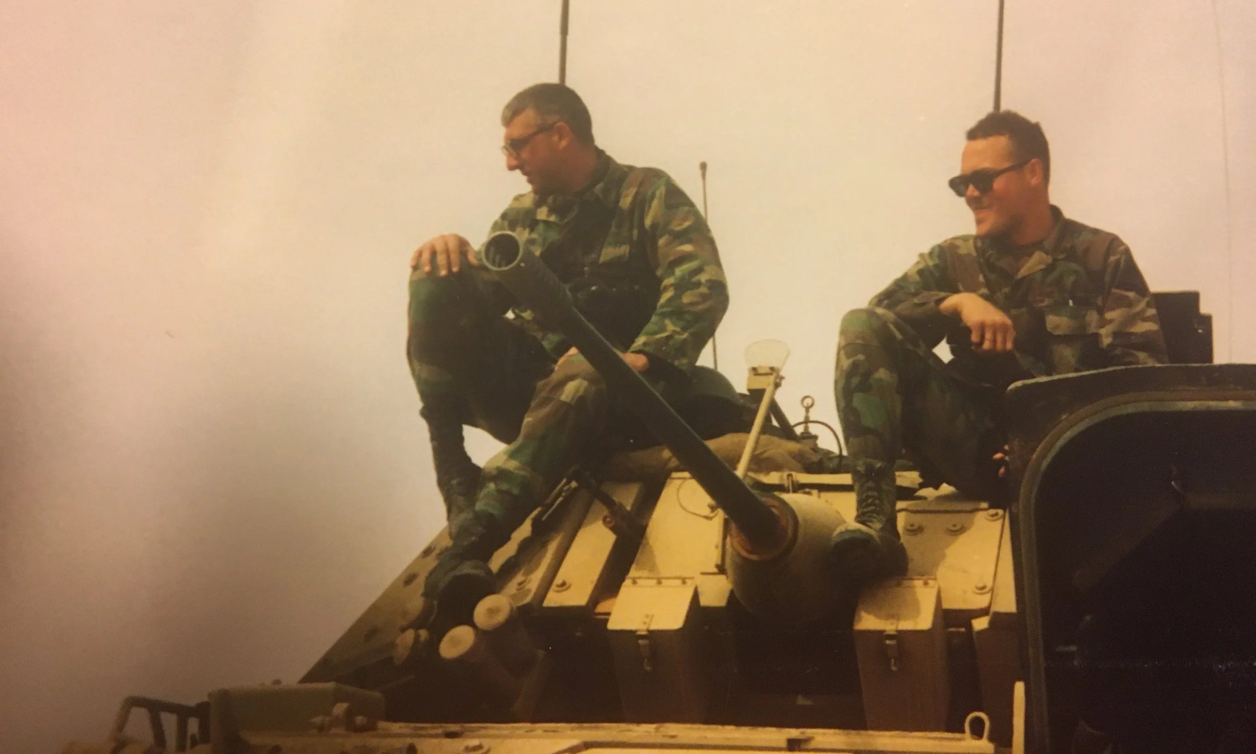 Two men in camouflage military uniforms sitting on a tank, smiling and relaxing.