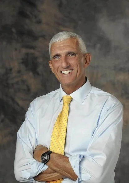 Mark Hertling smiling, dressed in a white shirt and yellow tie, standing in front of a dark, textured background.