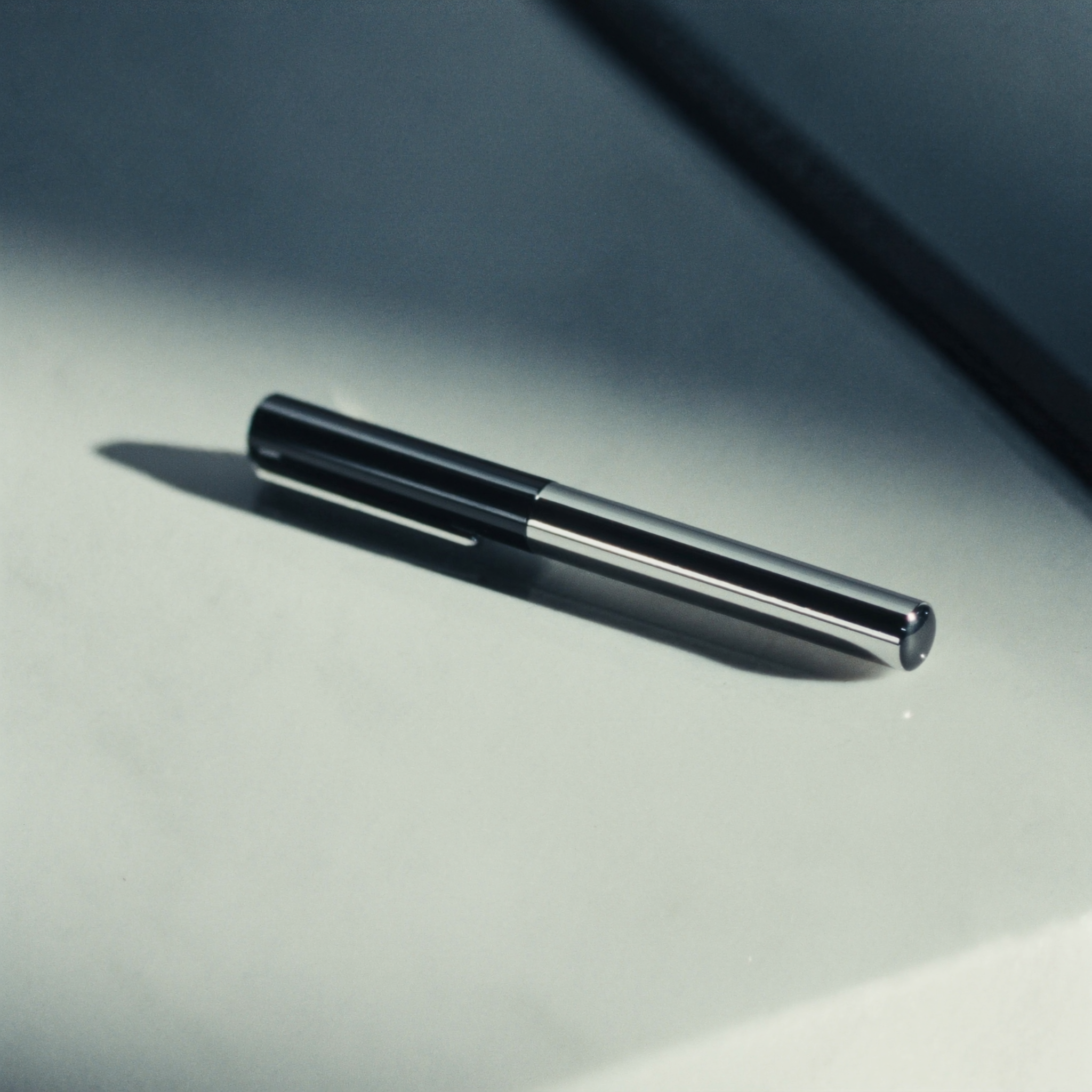 Close-up of a black ballpoint pen with a metallic clip casting a shadow on a light gray surface, with a dark blurred background.
