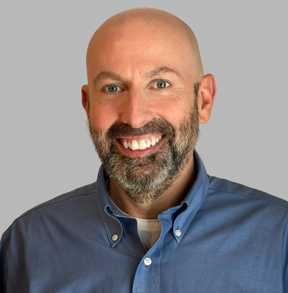 John smiling man with a bald head and gray beard, wearing a blue collared shirt, against a plain gray background.
