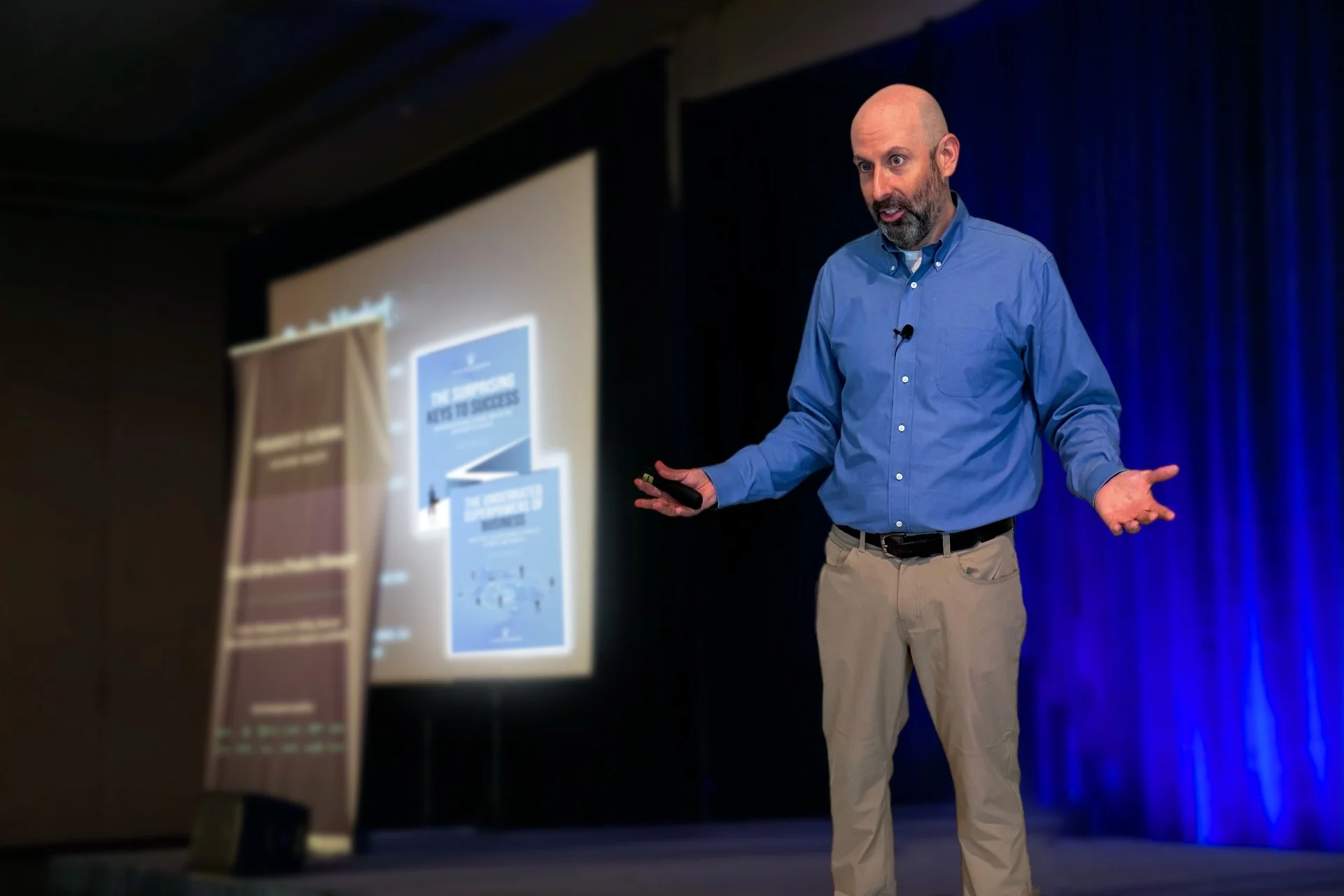 John in a blue button-up shirt and khakis giving a presentation on a stage with a blue curtain backdrop, gesturing with his hands.