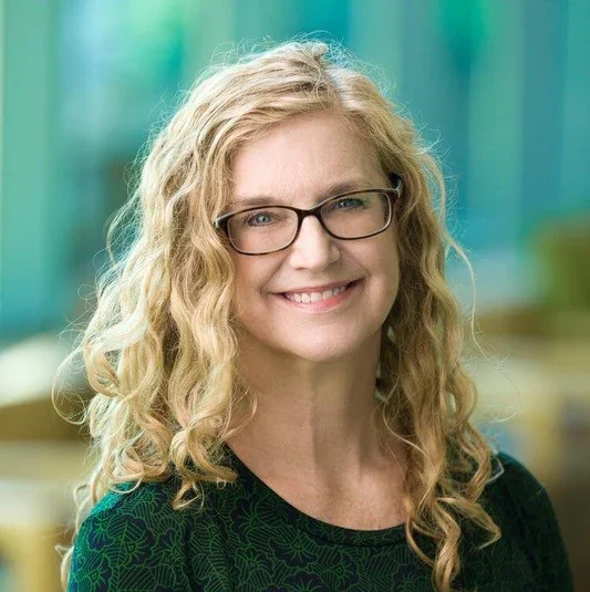 Headshot of a woman with curly blonde hair and glasses, smiling, indoors with a blurred background.