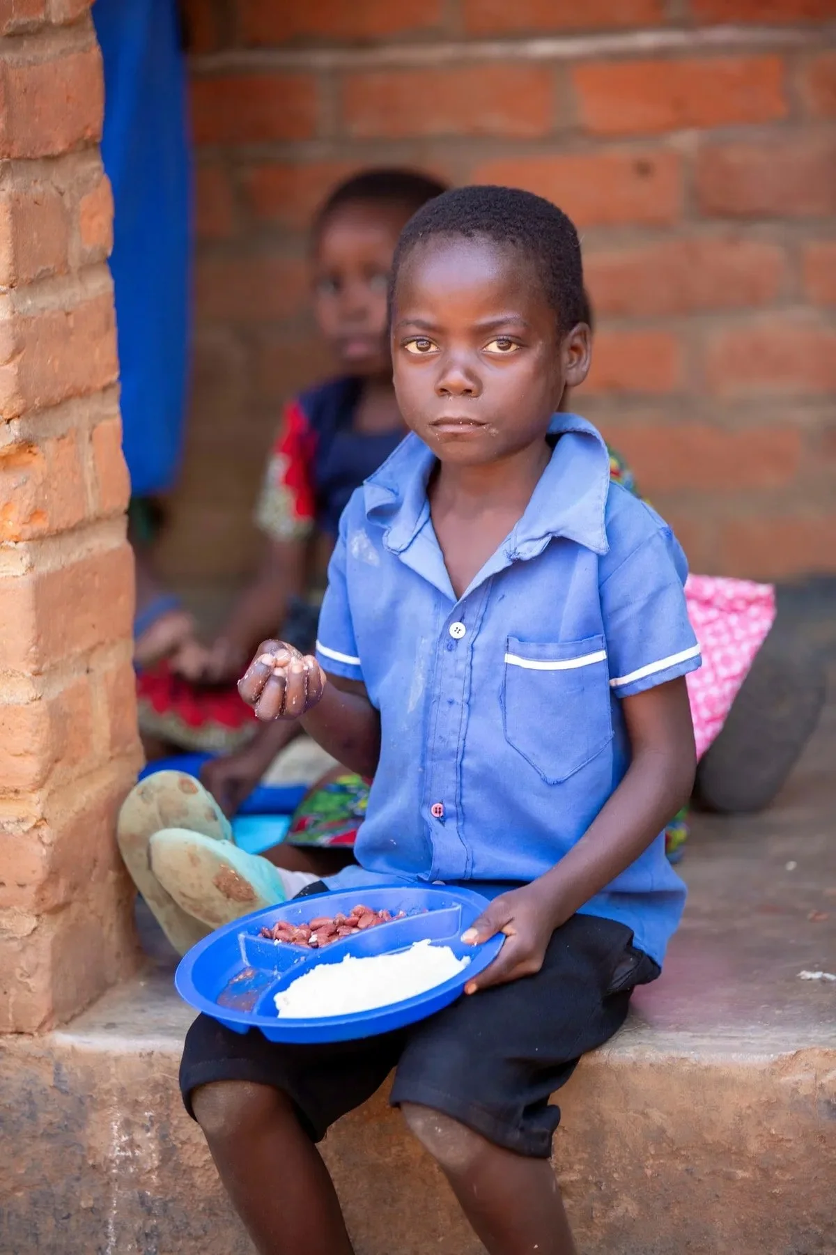 Young boy in a blue shirt sitting outside, holding a divided plate with food, with two children in the background against a brick wall.