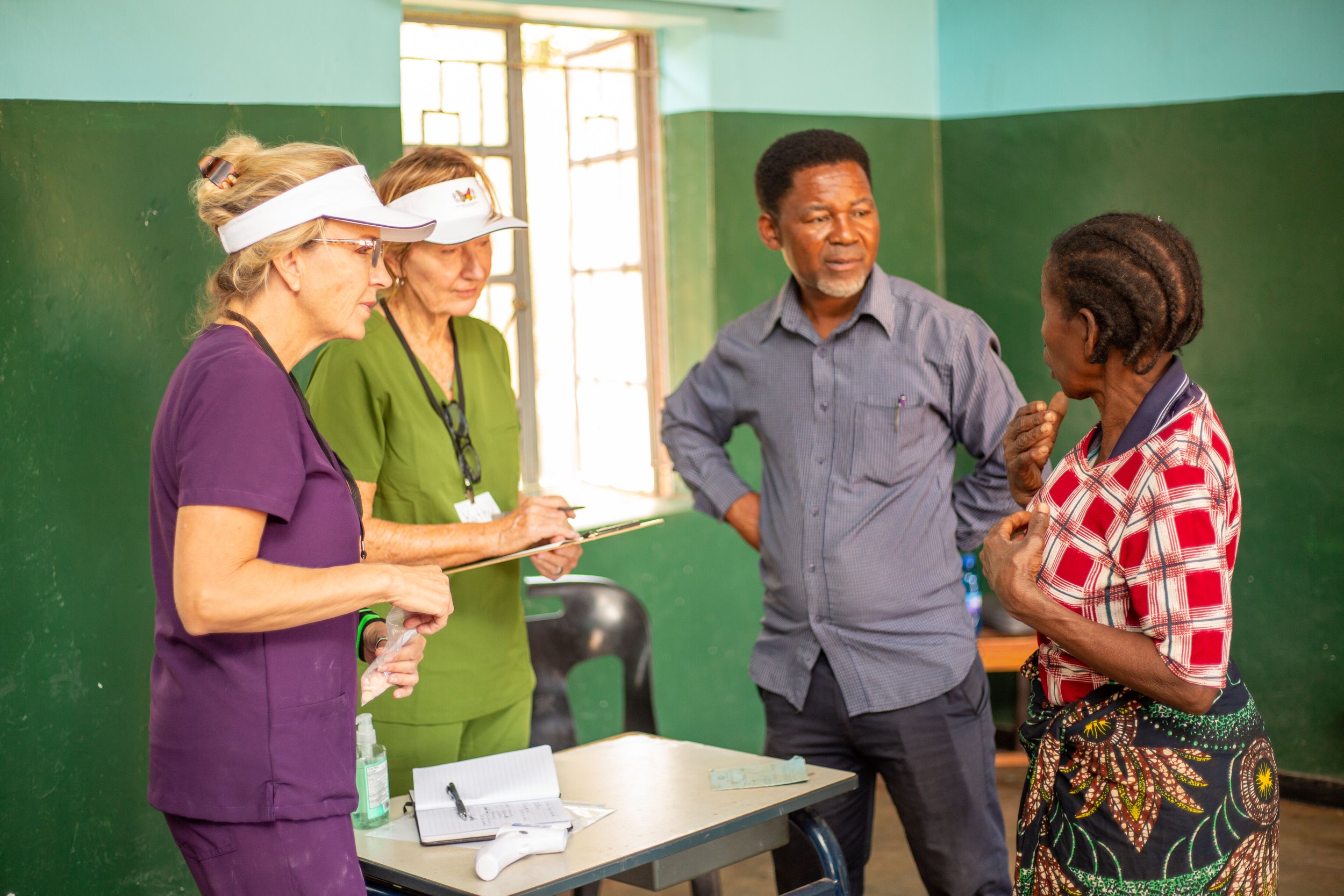 Group of four people engaged in conversation indoors, one woman in a red plaid shirt with short hair, two women in hats and scrubs, and a man in a gray shirt and short hair, with a green painted wall in the background.