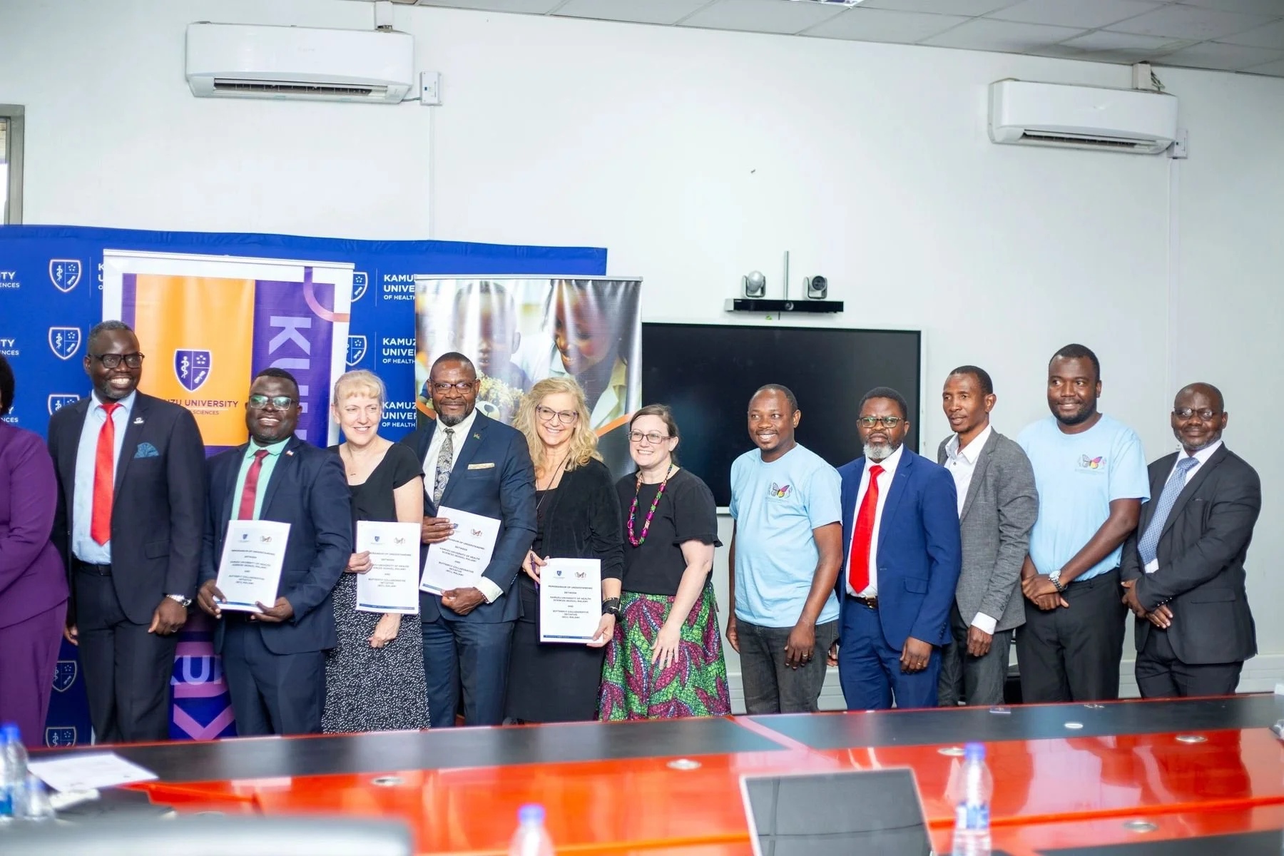 Group of diverse people in formal and casual attire standing in a row, some holding certificates, during a formal event at a university, with banners and a large screen in the background.
