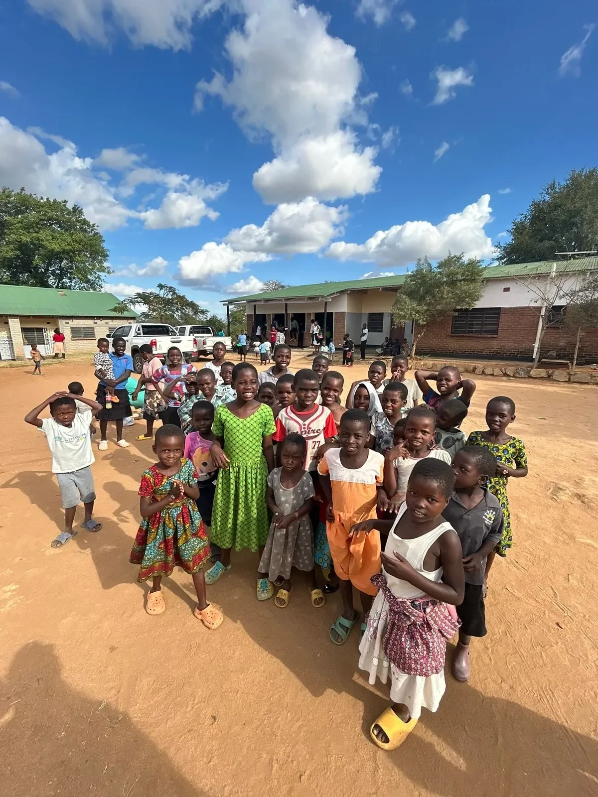Group of children and adults gathered outside a building on a sunny day with blue sky and clouds.