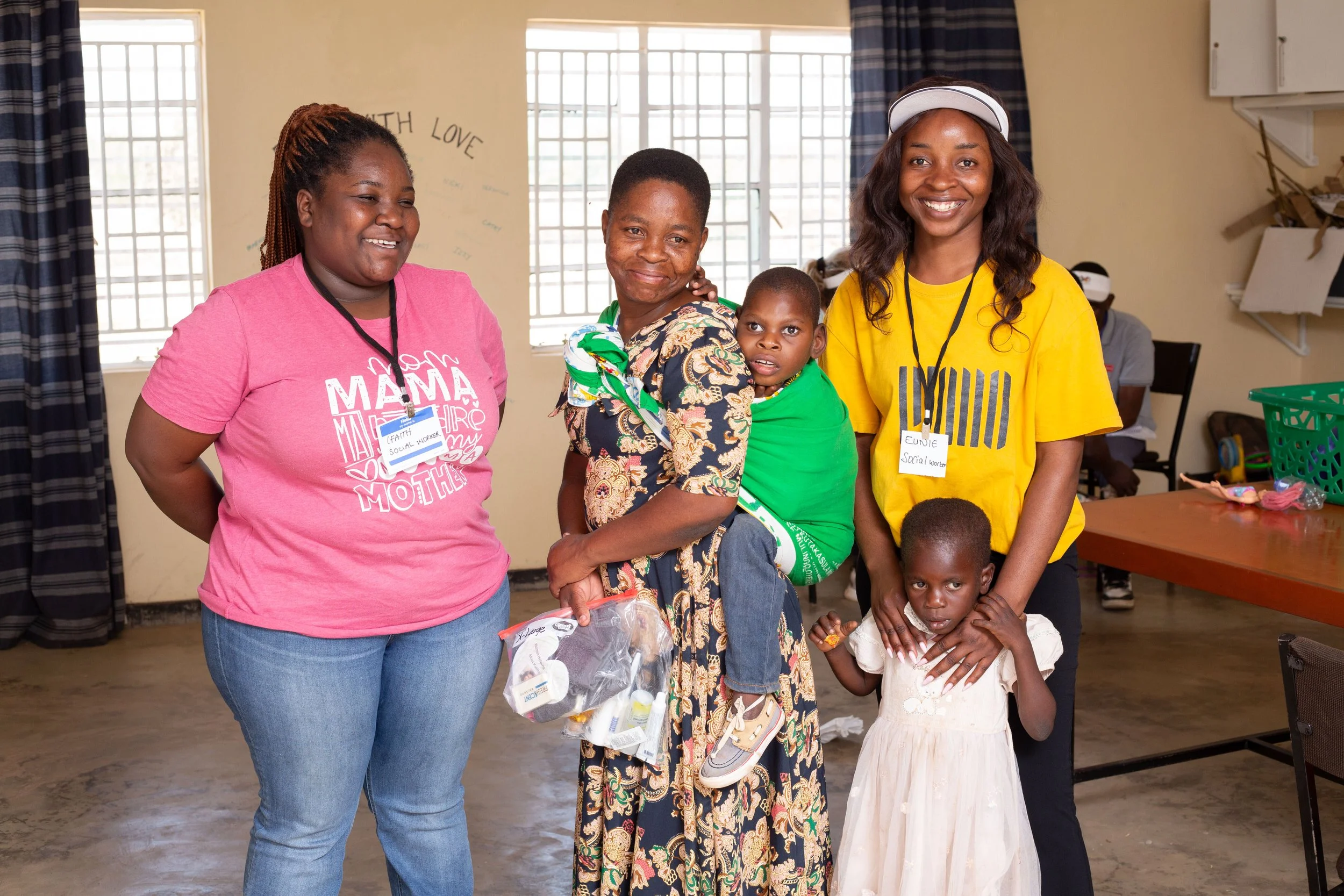 Group of smiling women and children in a room, some wearing name tags indicating they are social workers, with additional children and supplies in the background.