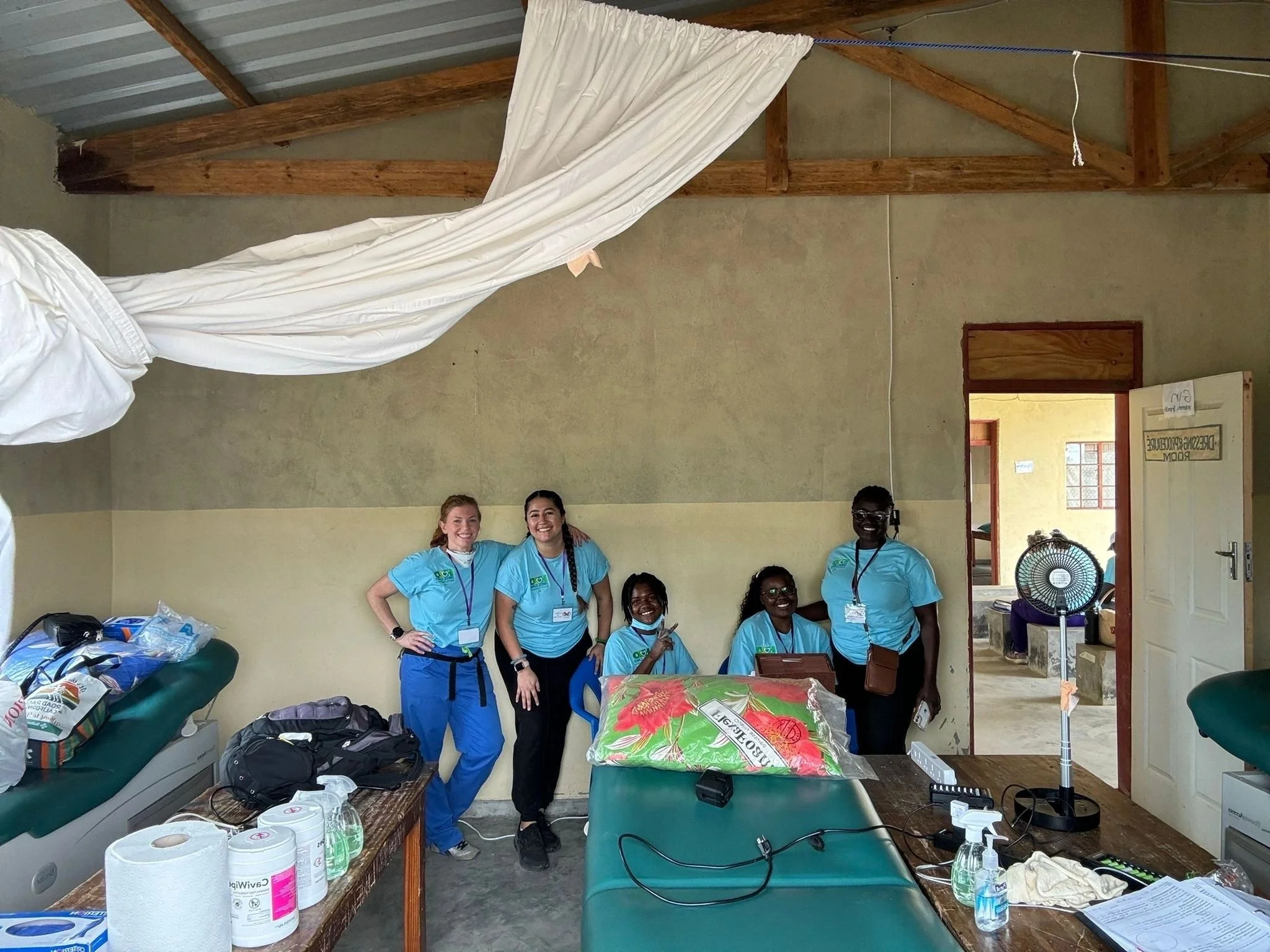 Group of smiling women in matching blue scrubs standing together inside a room, with supplies and a table in front of them. The room has a wooden ceiling and cream-colored walls.