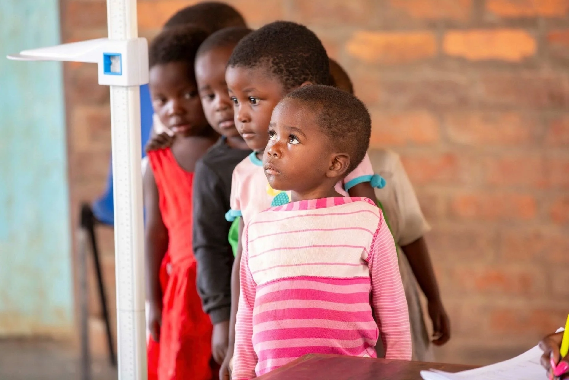 A line of five young children standing in a row inside a classroom, looking towards the left. The children are standing near a height measurement scale, with a brick wall in the background.