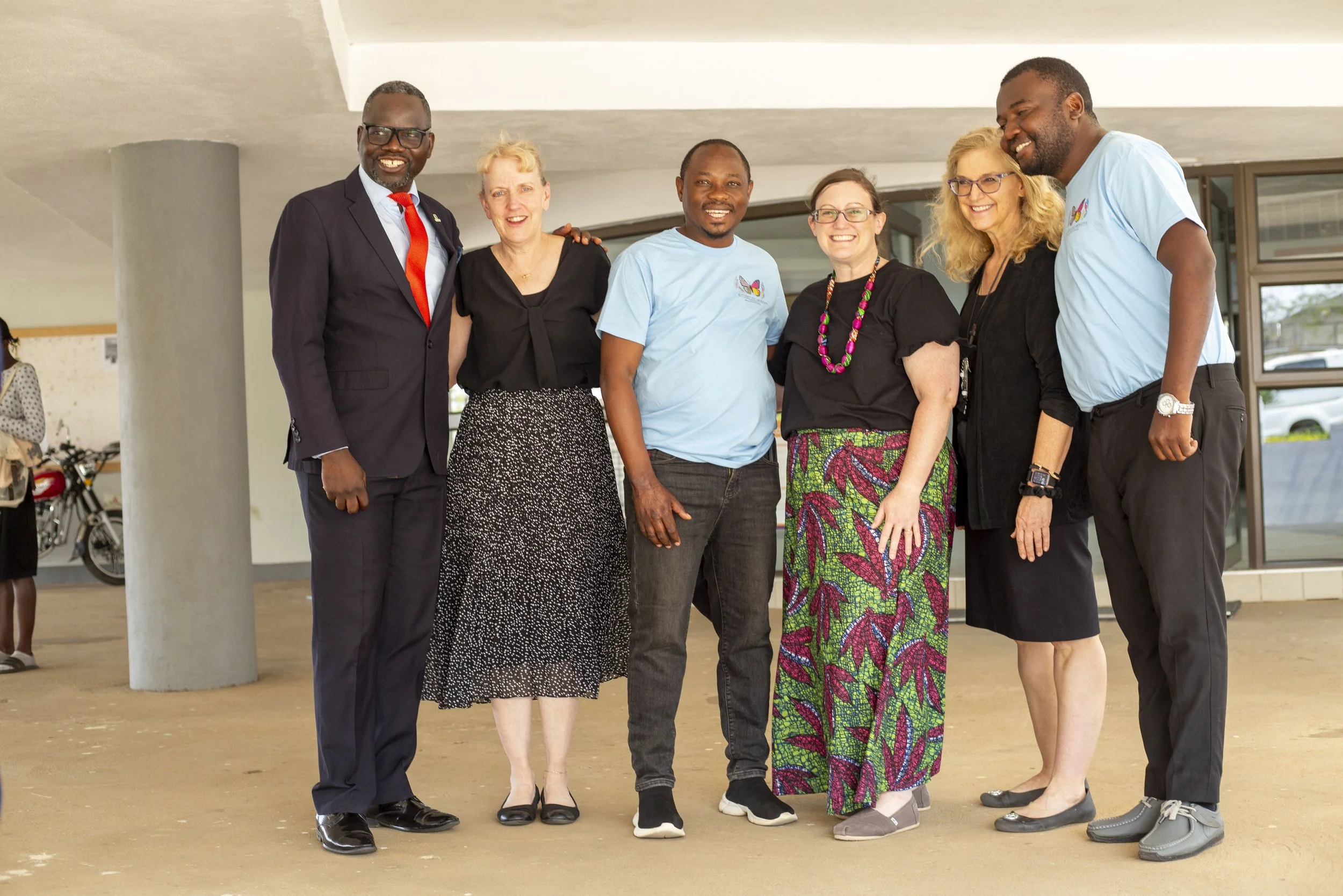 Group of six diverse people standing in a row outside, smiling for the photo. They are dressed in a mix of business casual and casual clothing, with a building entrance and windows visible in the background.