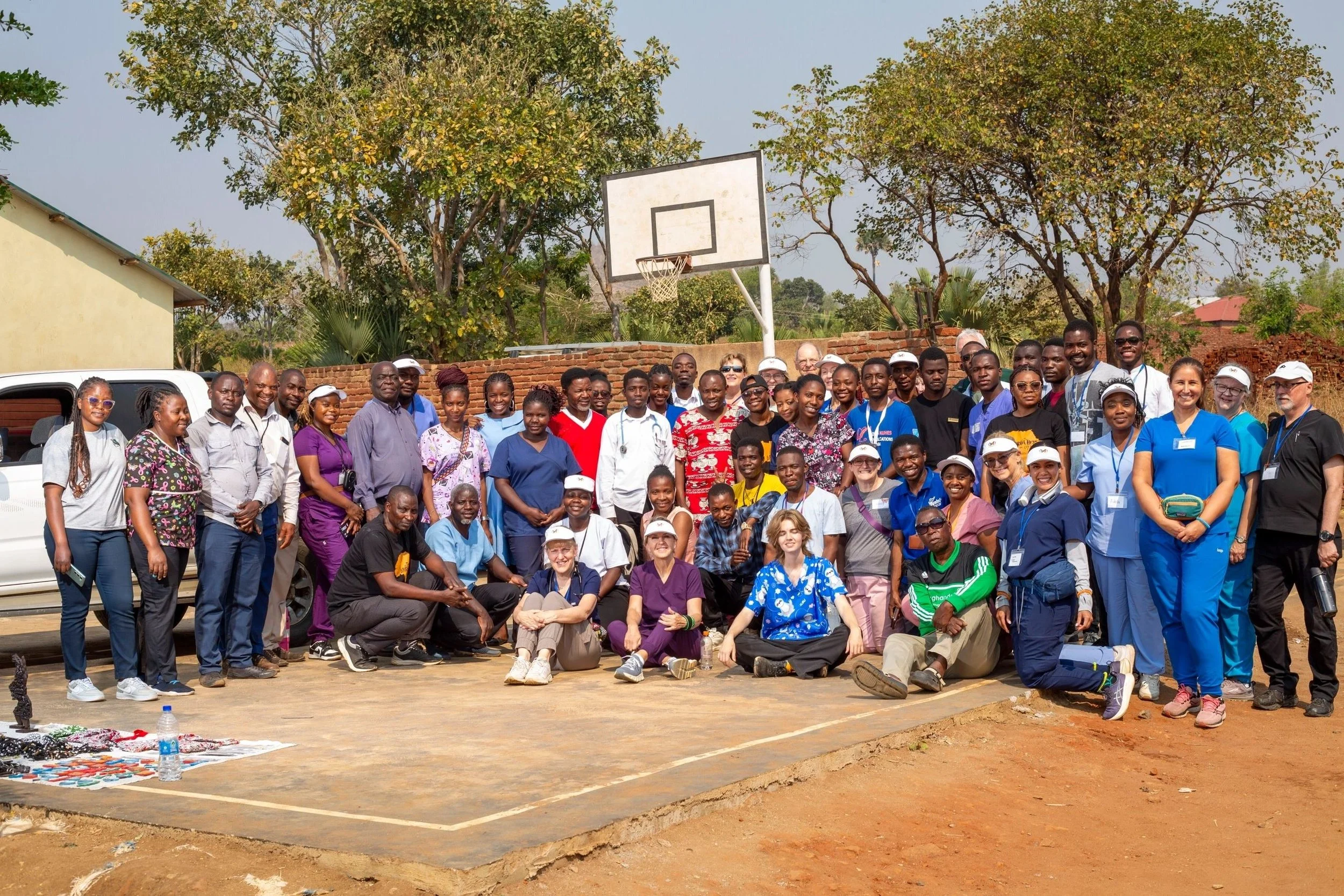 Group of diverse people standing on a soccer court outdoors, with a basketball hoop in the background, trees, and some buildings visible. The group is smiling and posing for a photo.