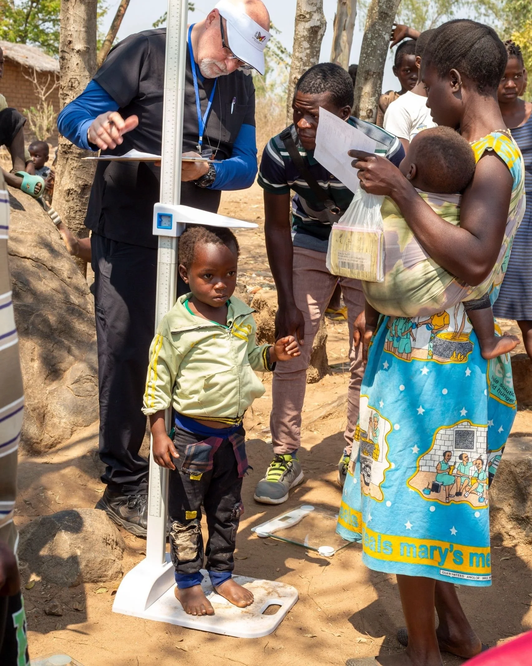 A man standing barefoot on a scale, being weighed by people around him, including a woman holding a child, in an outdoor setting with trees and a dirt ground.