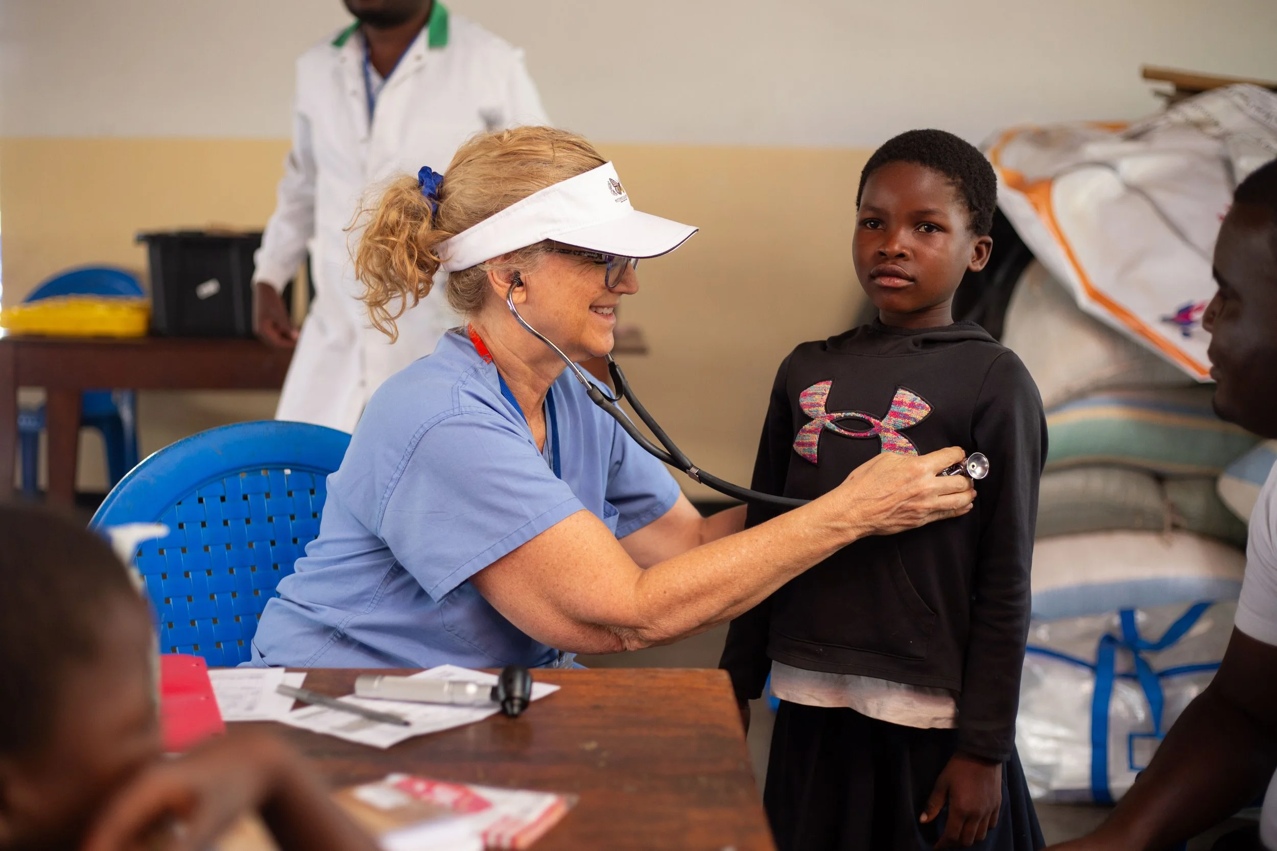 A healthcare professional in blue scrubs and a white visor examining a young girl with a stethoscope in a clinic or medical camp setting.