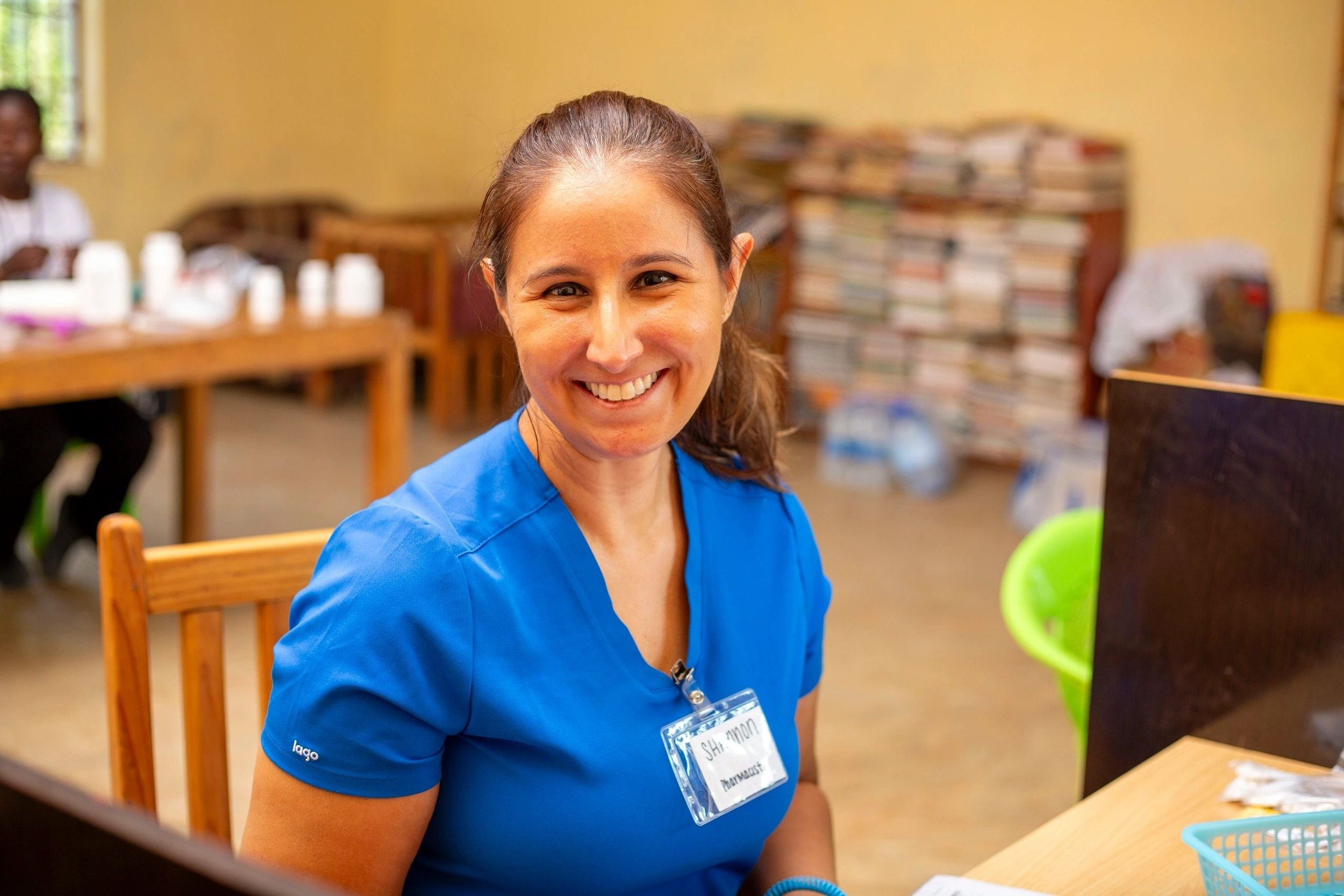 A woman smiling, wearing a blue shirt with a name tag, in a room with bookshelves and other people in the background.
