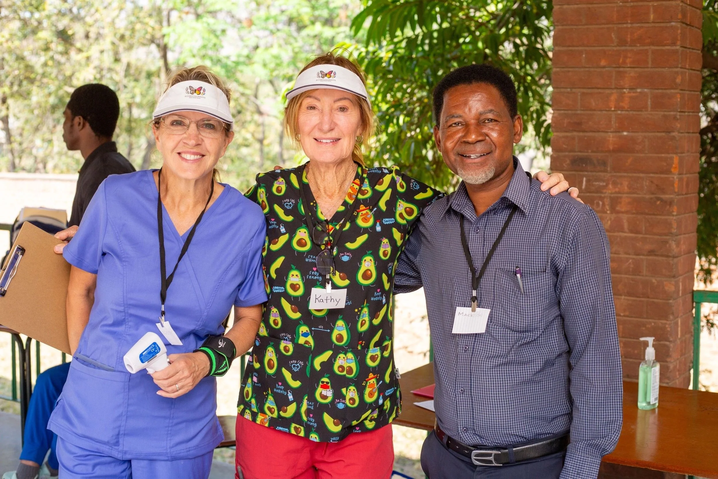 Three smiling people standing outdoors near a brick pillar, with a woman in medical scrubs holding a clipboard, and two older adults wearing colorful catering or event attire with name tags.