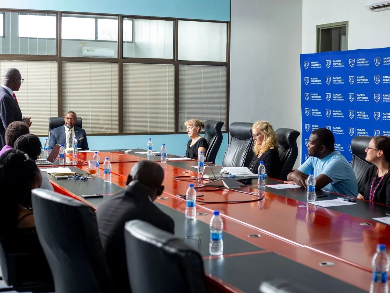 A diverse group of people attending a formal meeting or discussion around a large conference table in a modern office, with some taking notes and others listening attentively, in a room with large windows and a Kamuzu University of Health Sciences banner in the background.
