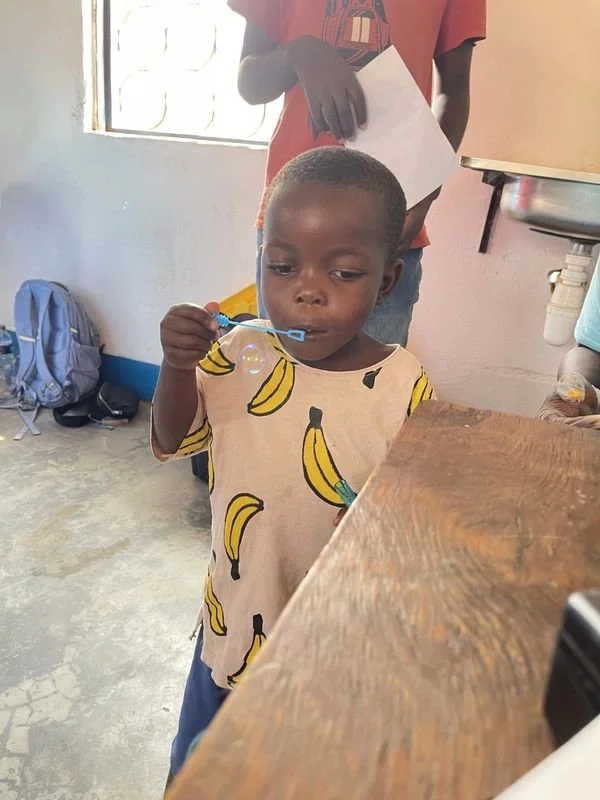 A young boy blowing bubbles with a bubble wand inside a room with a concrete floor, a window, and a sink. An adult is standing behind him holding a piece of paper. A backpack is on the floor in the background.