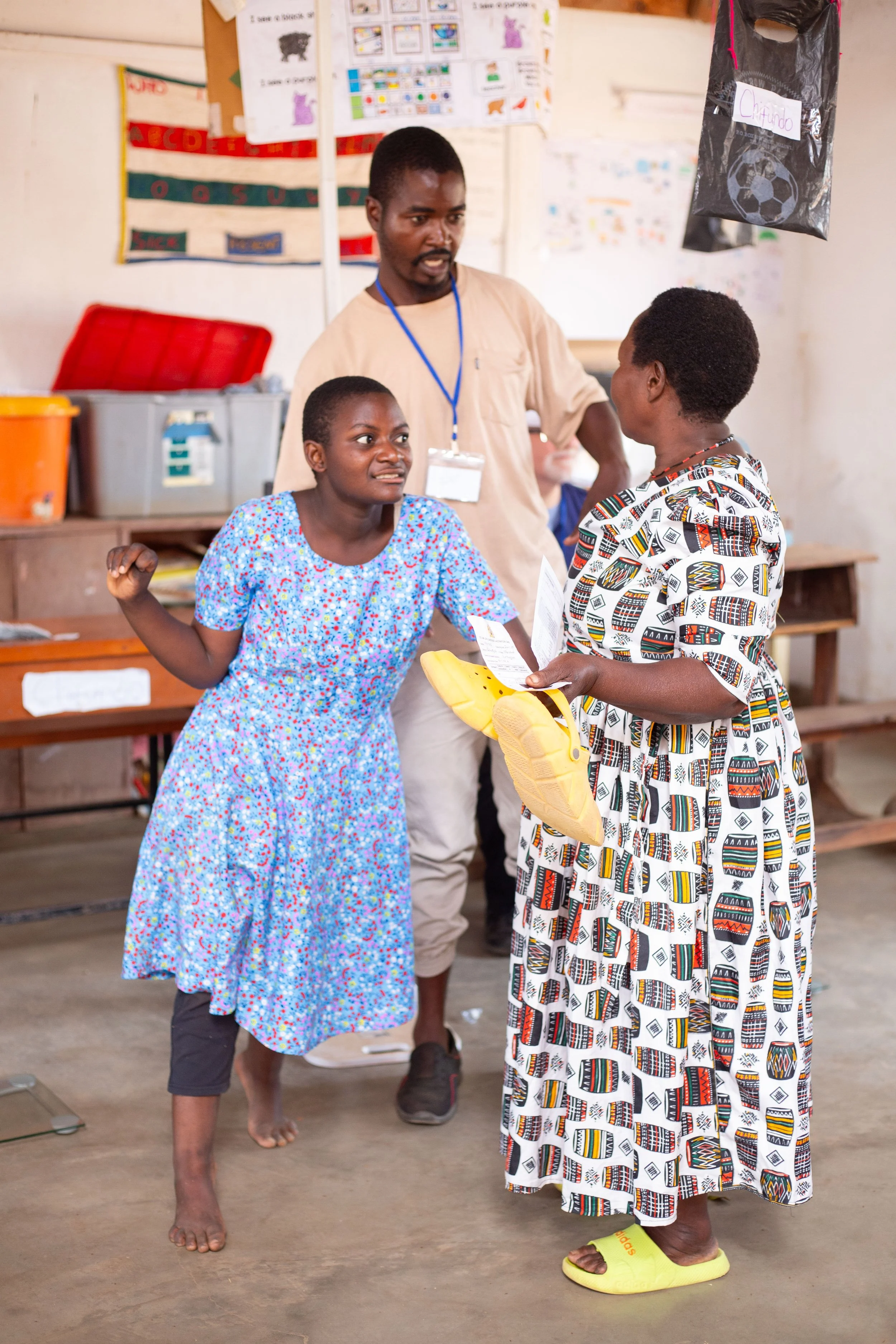 Two women and a man in a classroom engaging in a discussion. The woman on the left wears a blue floral dress, and the woman on the right wears a dress with a colorful geometric pattern. The man stands between them, wearing a beige shirt, eyeglasses, 