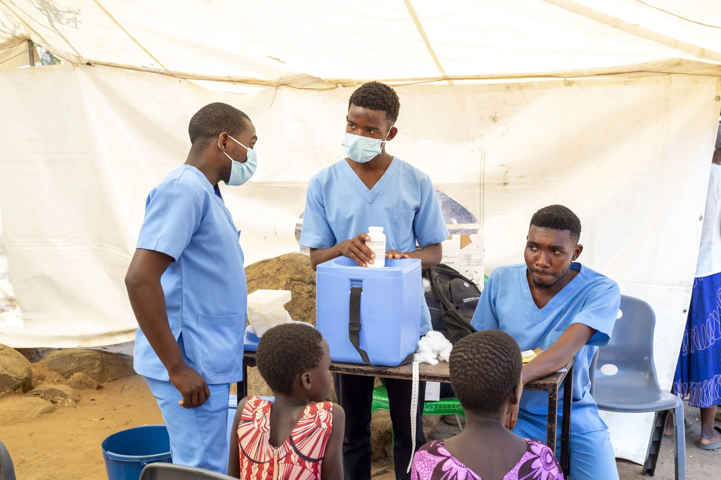 Medical professionals wearing masks and scrubs engaged in a discussion at an outdoor healthcare event, with children and a tent in the background.