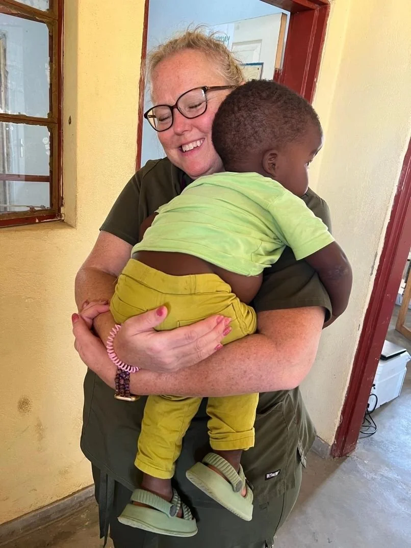 A woman with glasses and red hair hugging a small child. The woman is smiling with closed eyes, and the child, wearing a light green shirt and yellow pants, is resting against her chest. They are indoors near a window and a door, with a yellow wall i